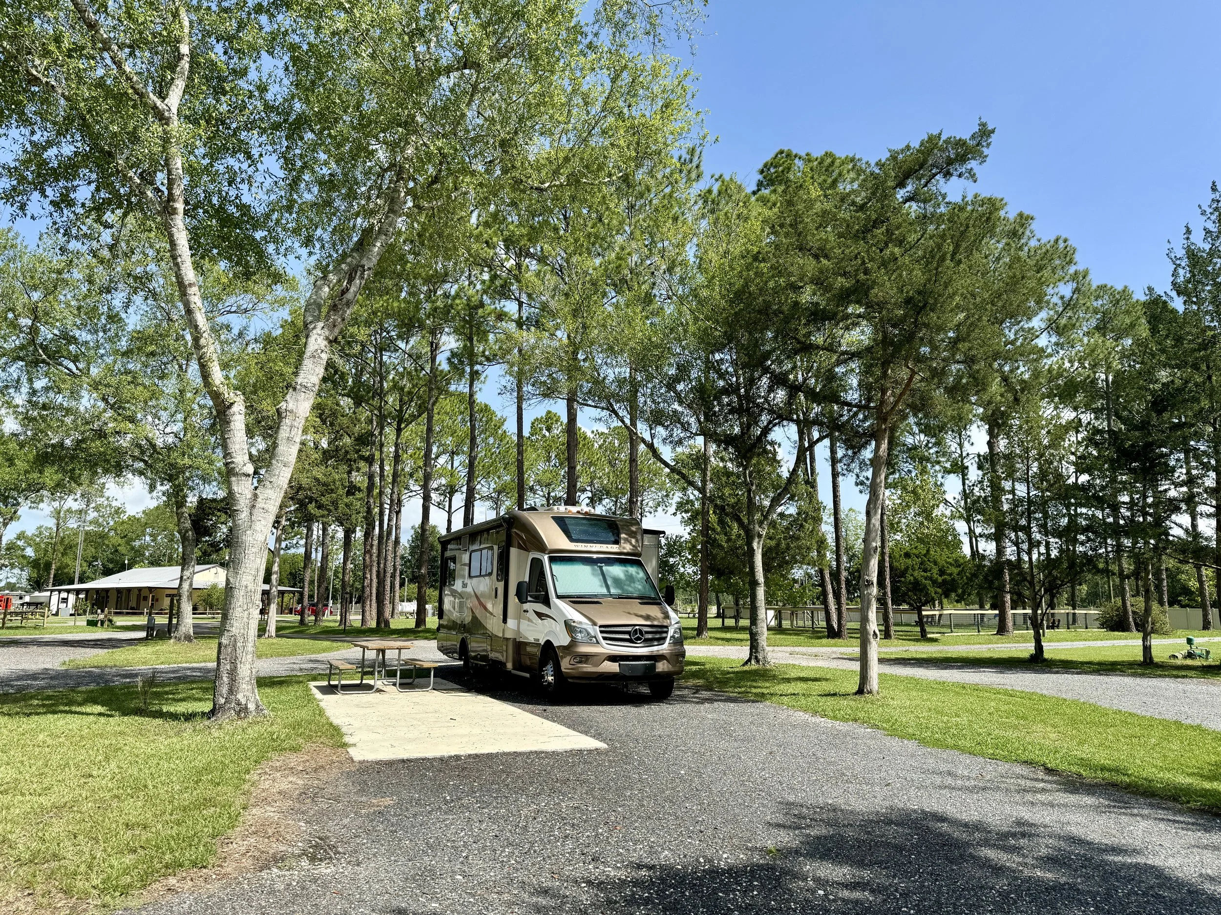 A recreational vehicle parked at a campsite surrounded by green trees and a gravel path, with a picnic table nearby under a clear blue sky.
