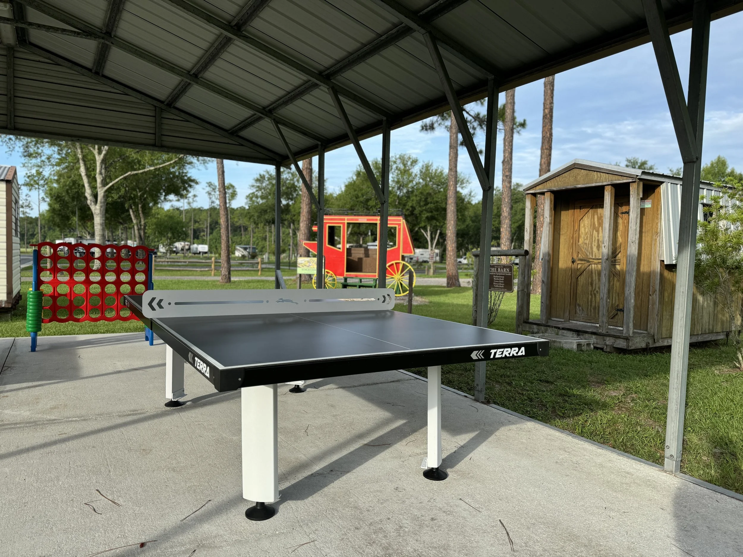 Outdoor covered area with a ping pong table, giant connect four game, and a small wooden shed, with trees and parked cars in the background.