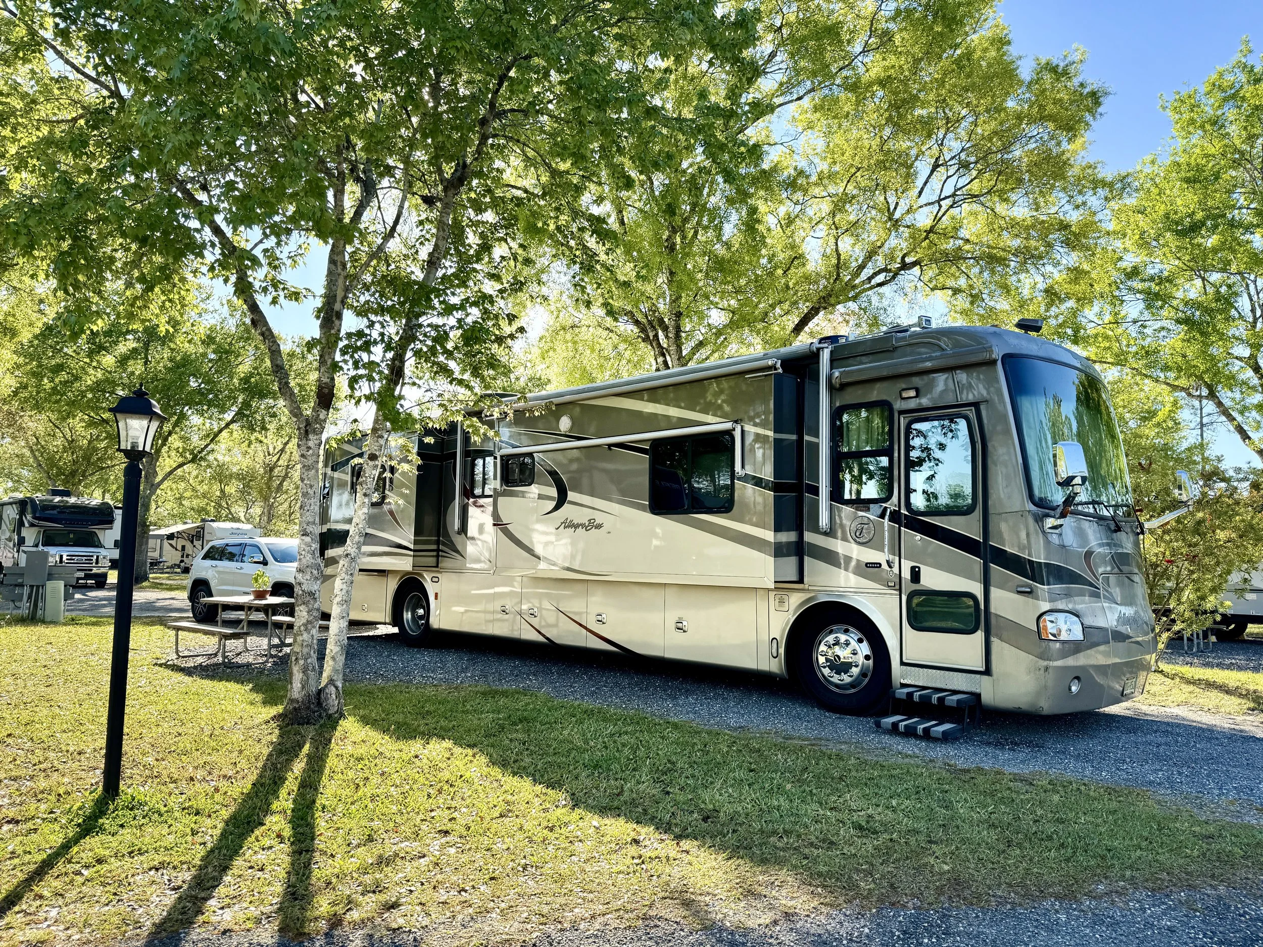 A large beige and gray RV parked on a gravel pad under green trees, with a picnic table and a black lamp post nearby in a campground.