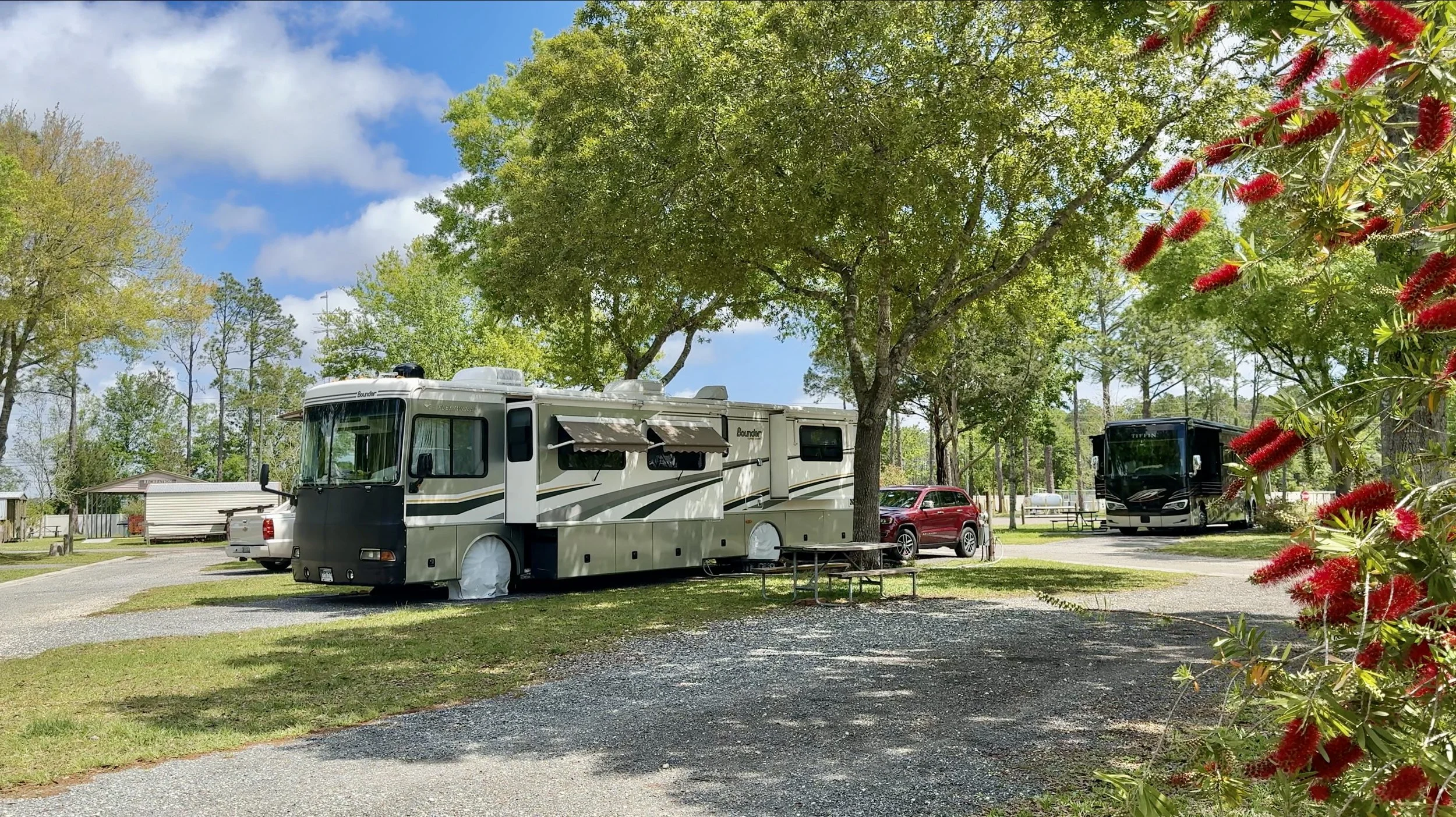 A recreational vehicle parked in a campground with green trees, a picnic table, and a blue sky with clouds.