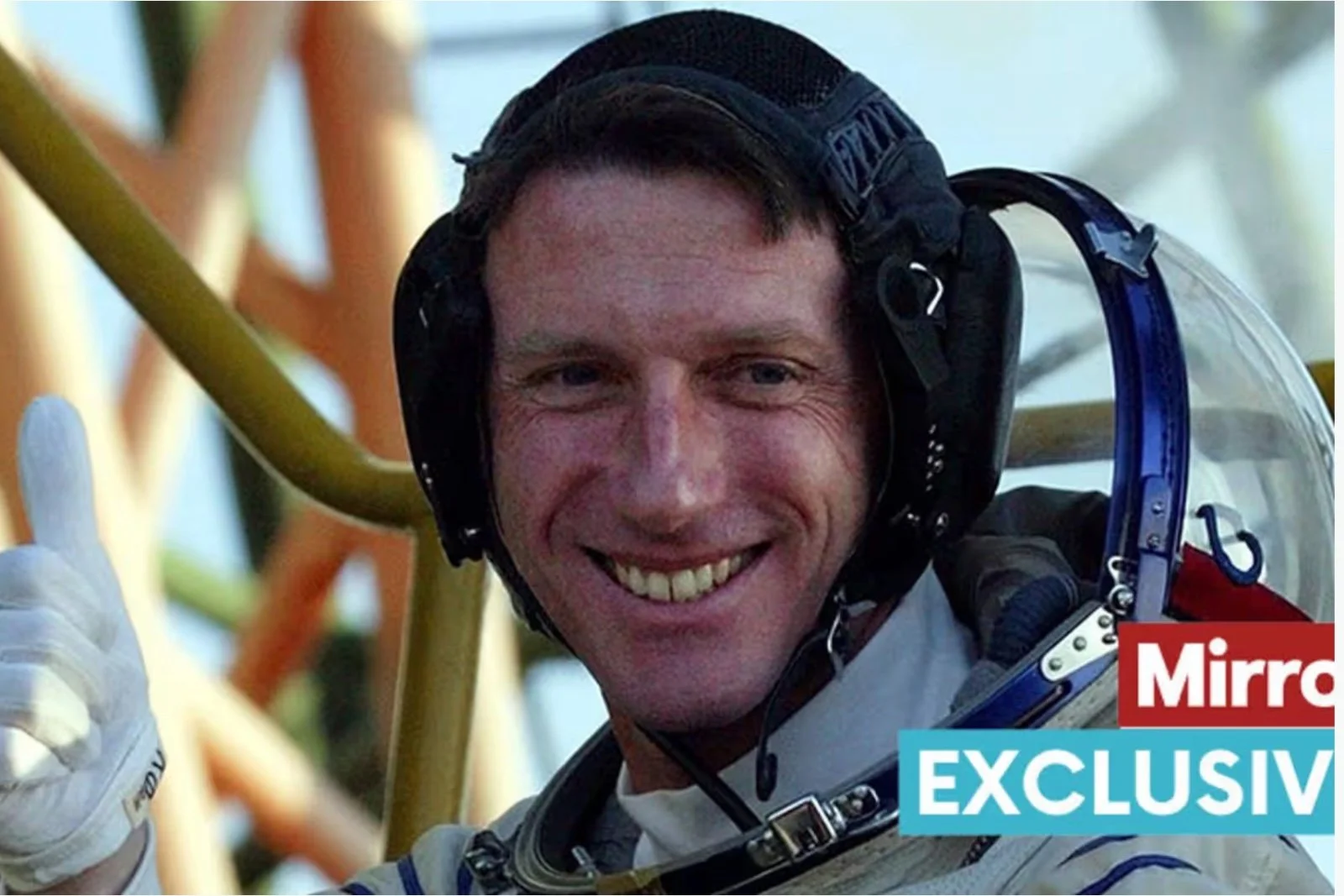 A smiling man in a spacesuit giving a thumbs up, with parts of a spacecraft visible in the background.