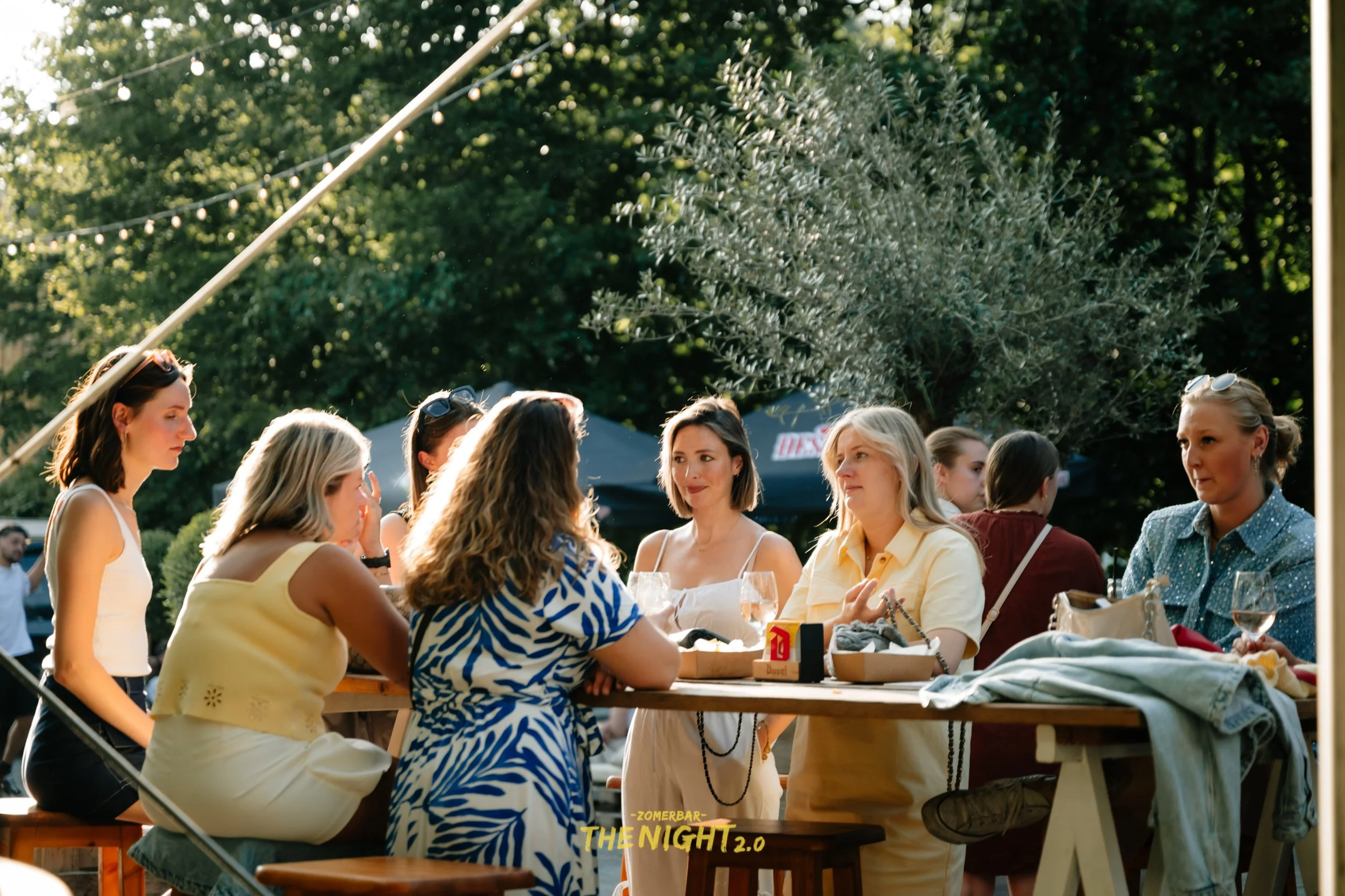 People dining at an outdoor bar or restaurant with string lights and wicker lamps hanging from the ceiling, green trees and umbrellas in the background.