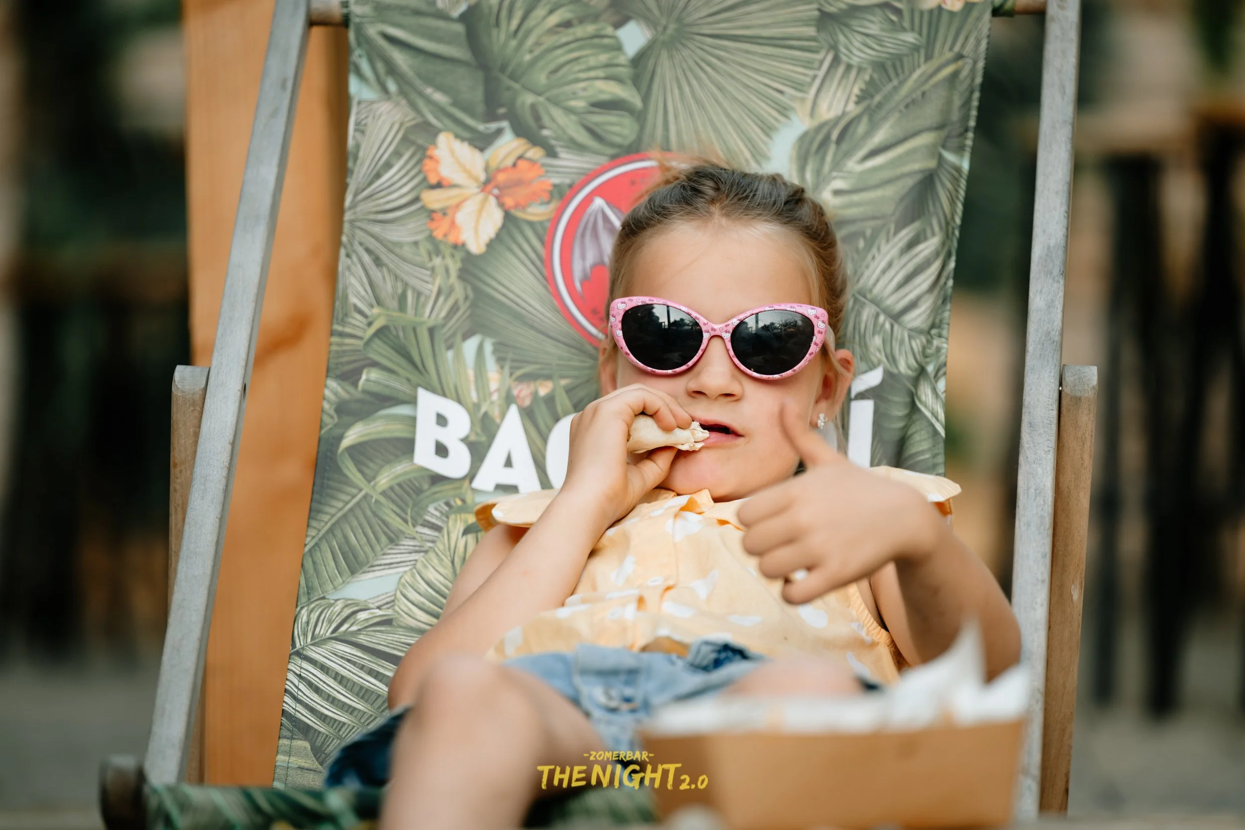 People dining at an outdoor bar or restaurant with string lights and wicker lamps hanging from the ceiling, green trees and umbrellas in the background.