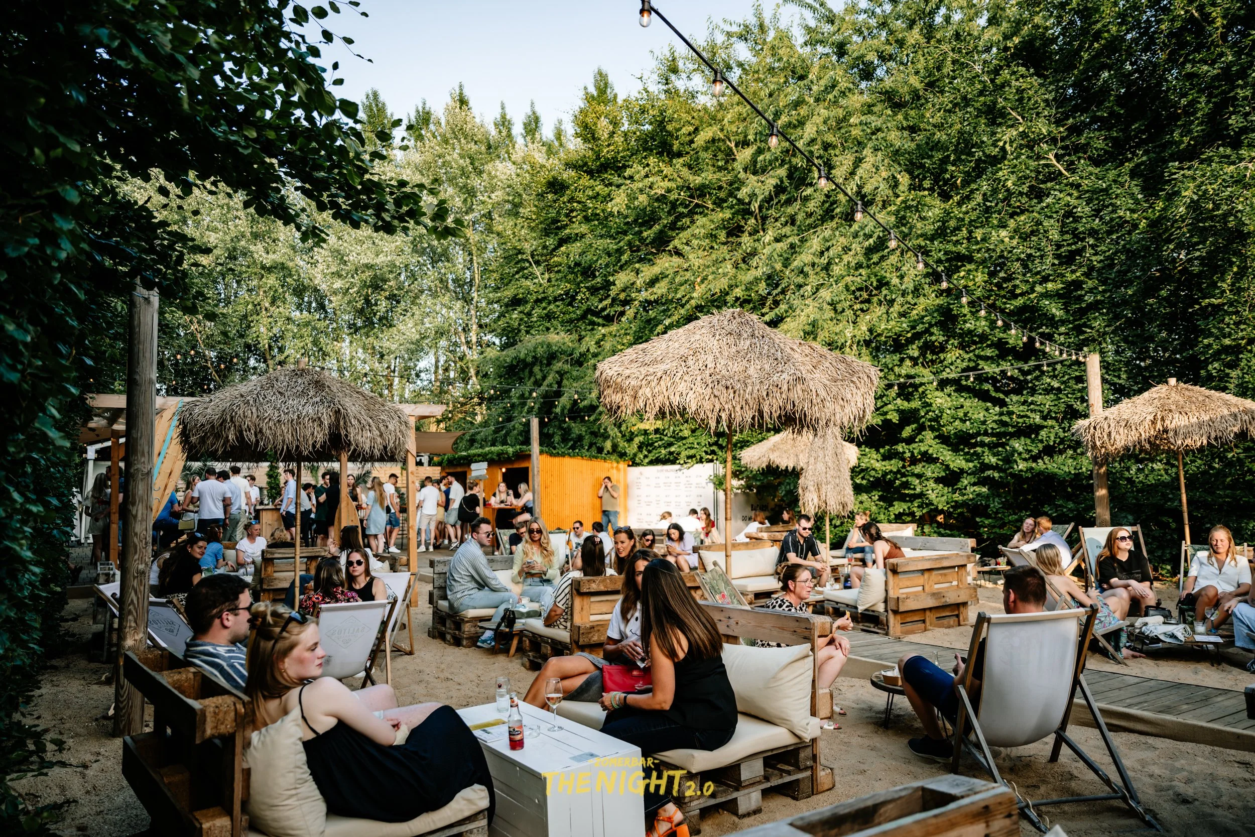 Outdoor dining area at sunset with string lights, umbrellas, and groups of people socializing and eating.