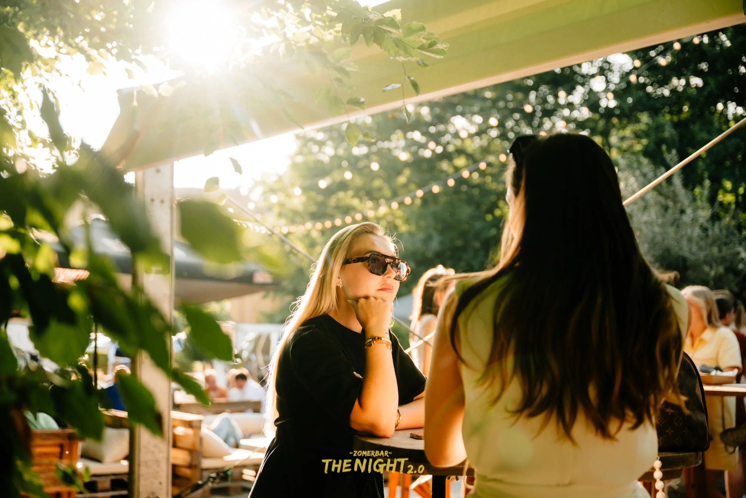 A group of people gathered at an outdoor bar or restaurant, surrounded by green trees, with sunlight filtering through the leaves, and the text "ZOMERBAR THE NIGHT 2.0" at the bottom.