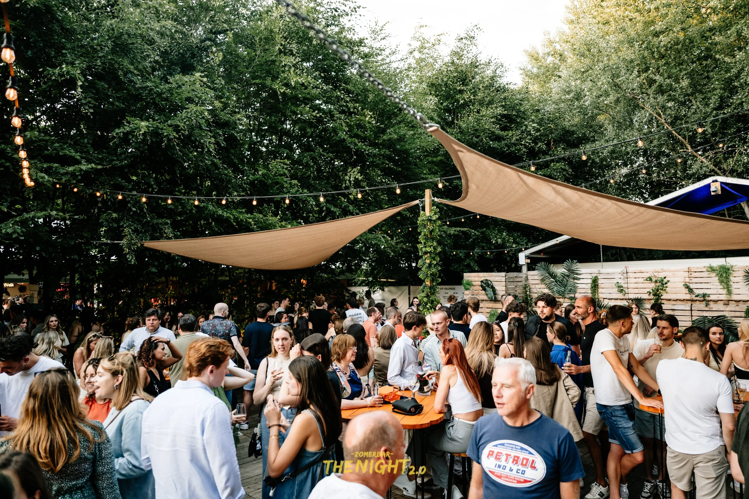 Outdoor dining area at sunset with string lights, umbrellas, and groups of people socializing and eating.
