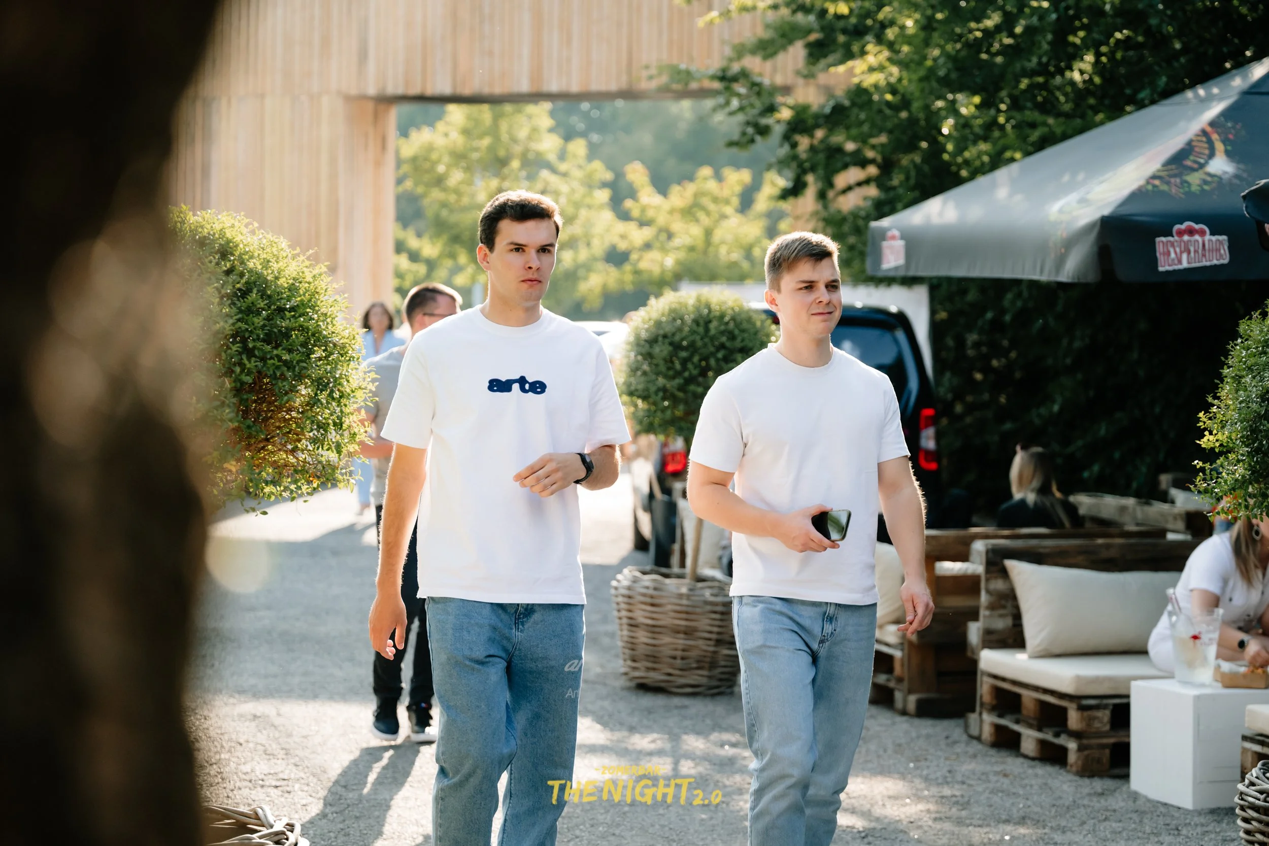 People dining at an outdoor bar or restaurant with string lights and wicker lamps hanging from the ceiling, green trees and umbrellas in the background.