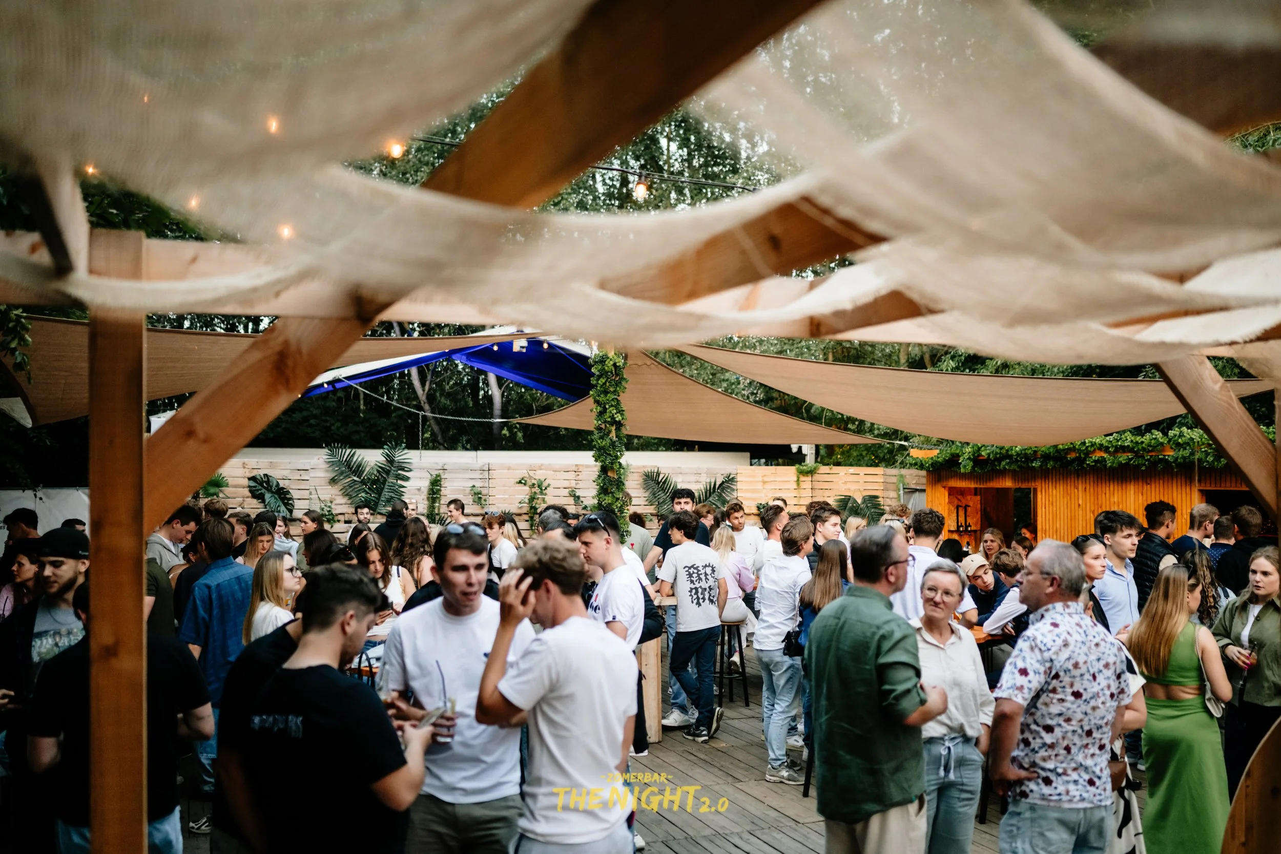 Outdoor dining area at sunset with string lights, umbrellas, and groups of people socializing and eating.