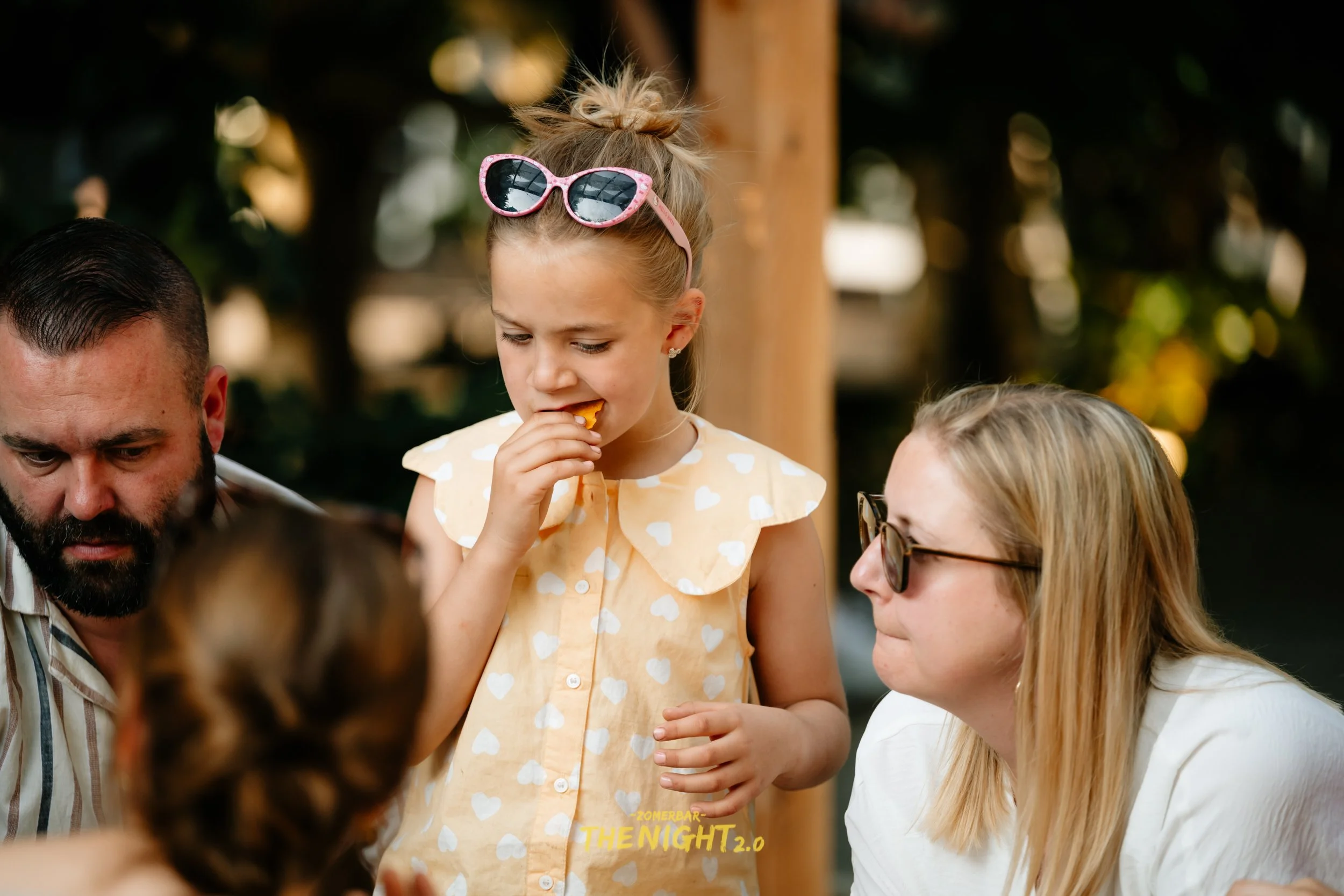 People dining at an outdoor bar or restaurant with string lights and wicker lamps hanging from the ceiling, green trees and umbrellas in the background.