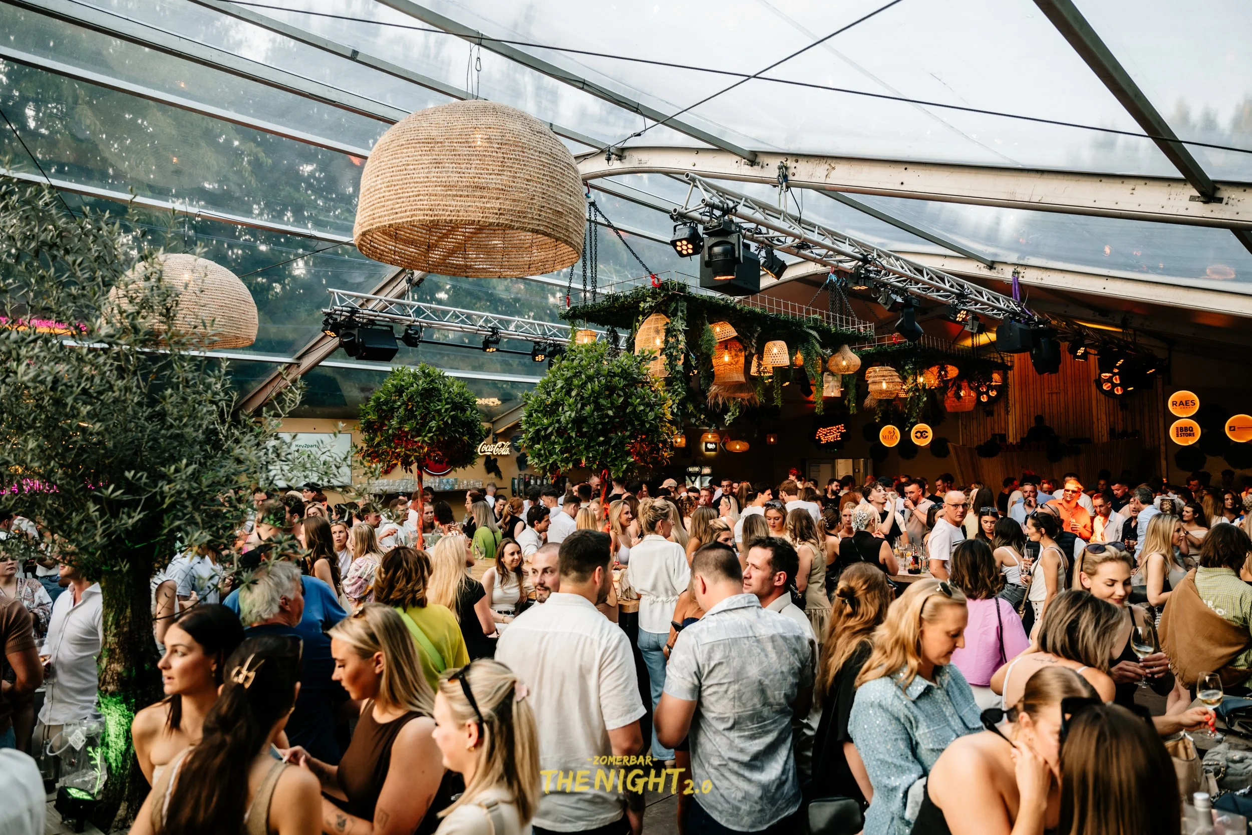 Crowded indoor event with numerous people sitting at tables and standing, decorated with hanging lanterns and greenery.