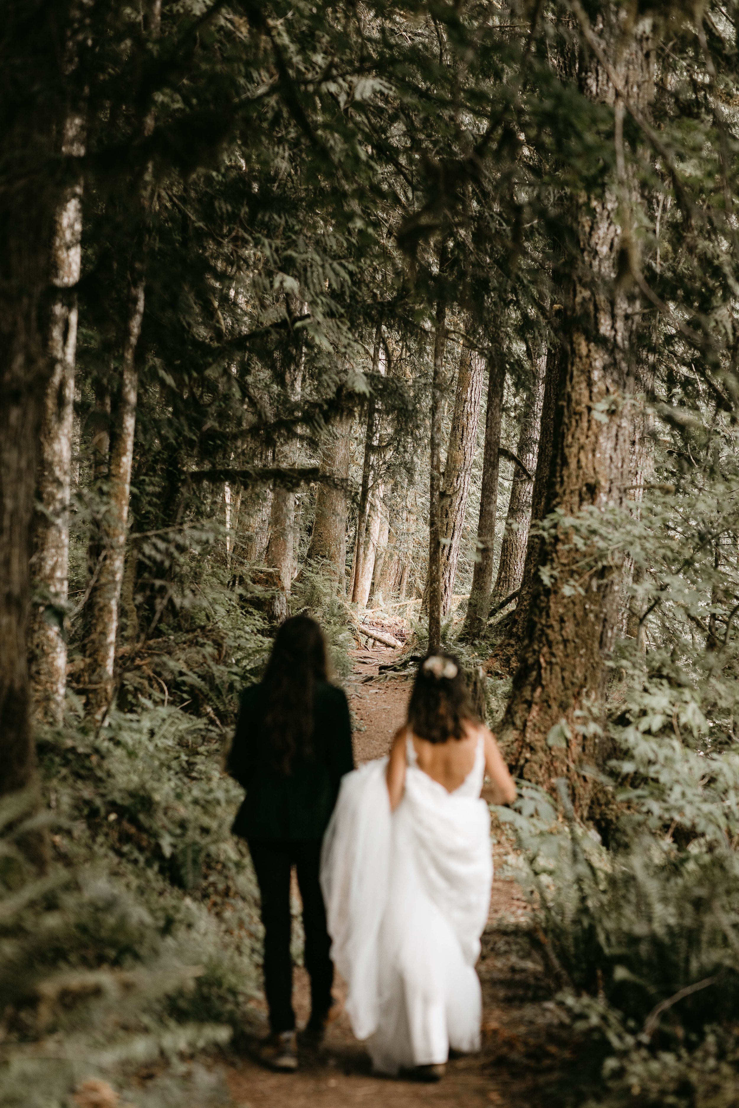  A couple walks down a trail in the North Cascades during their elopement at Artist Point. One woman is wearing a dark suit and the other a wedding gown. 