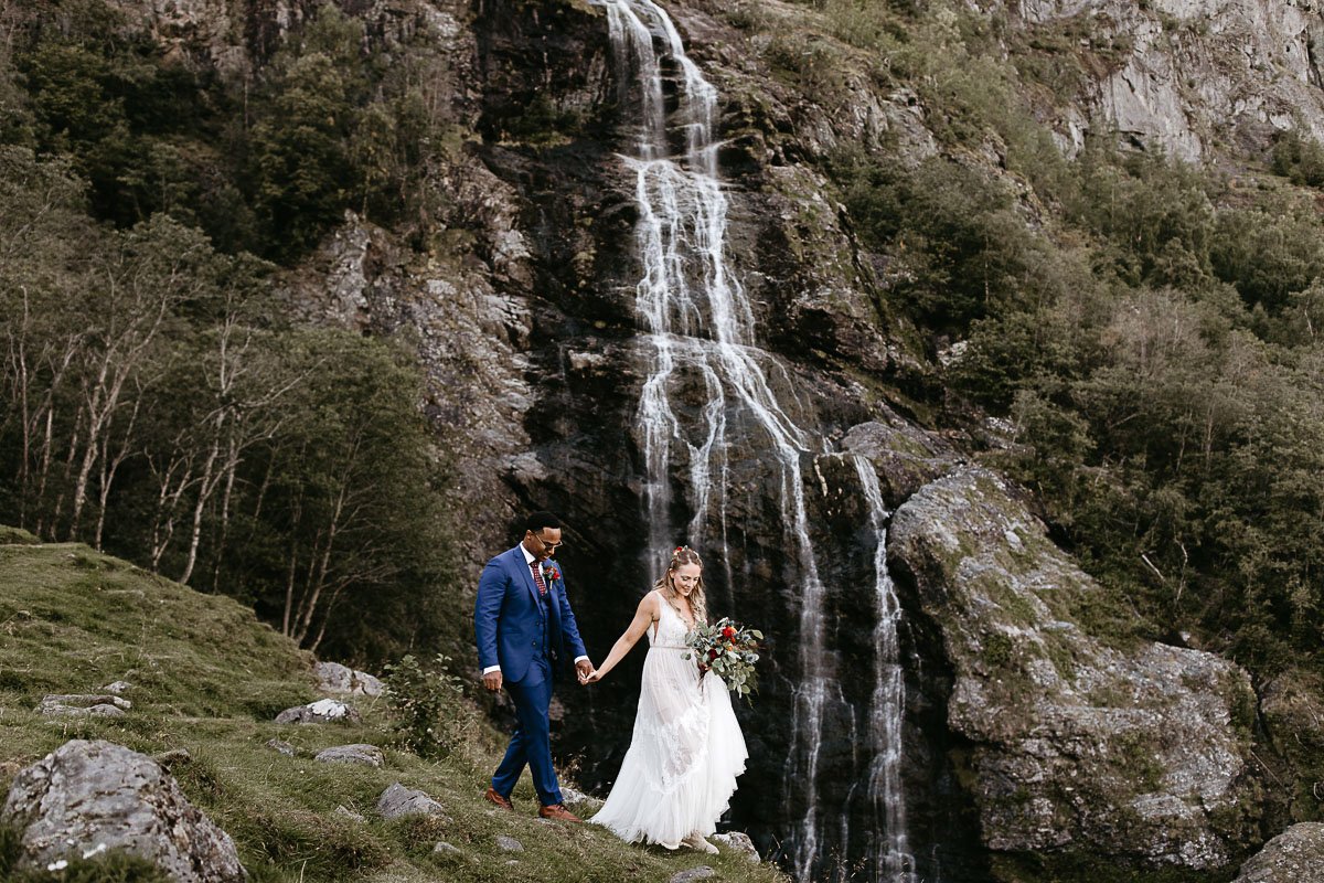  This is an image of a couple eloping under a waterfall in Flåm, Norway. They are wearing wedding attire and standing next to the water. 