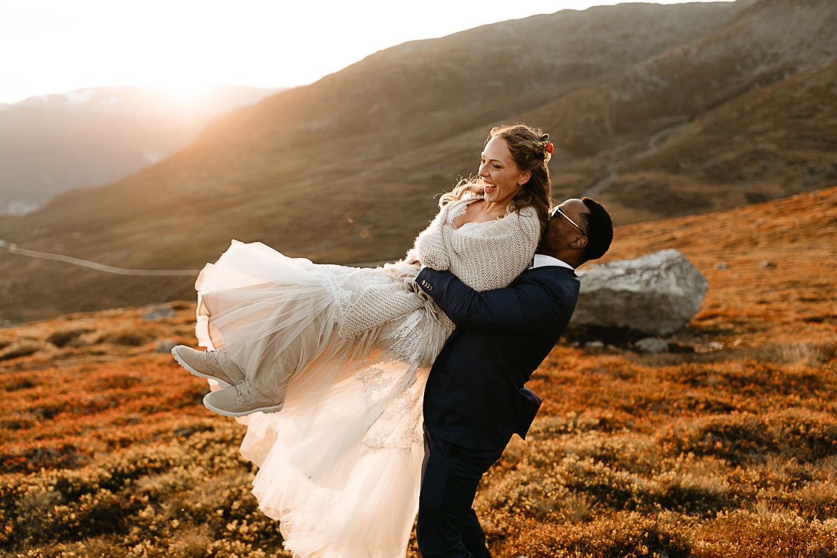  This is a photo of a couple eloping near Flåm Norway. They are in a field full of grasses and the sun is a golden yellow. The man is wearing a dark navy suit and picking up the bride, who is laughing and wearing a long white dress and sweater. 