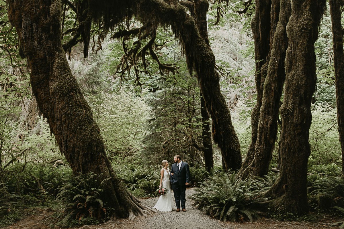 A couple stands in the Hoh Rainforest surrounded by towering, mossy trees, wearing their elopement clothes  