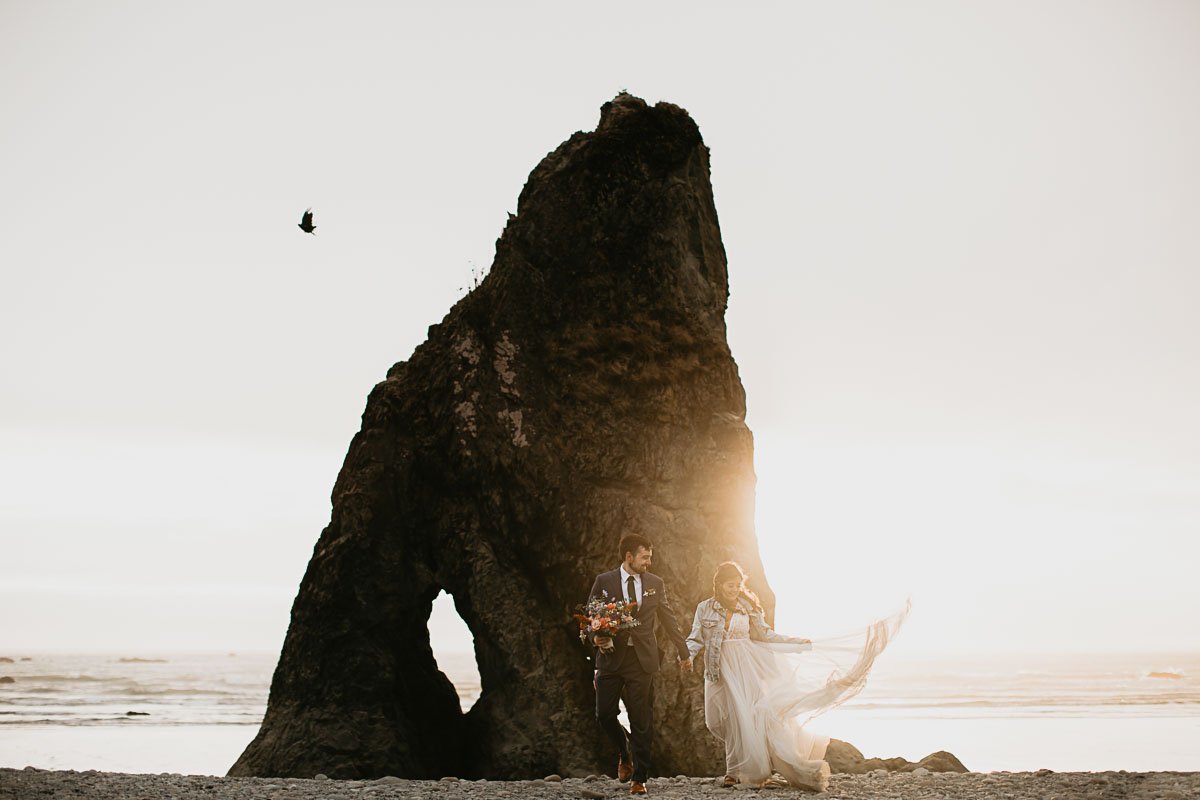  A couple stands on Ruby beach in their wedding attire looking at the sand. A large sea stack frames the sun setting behind them. 