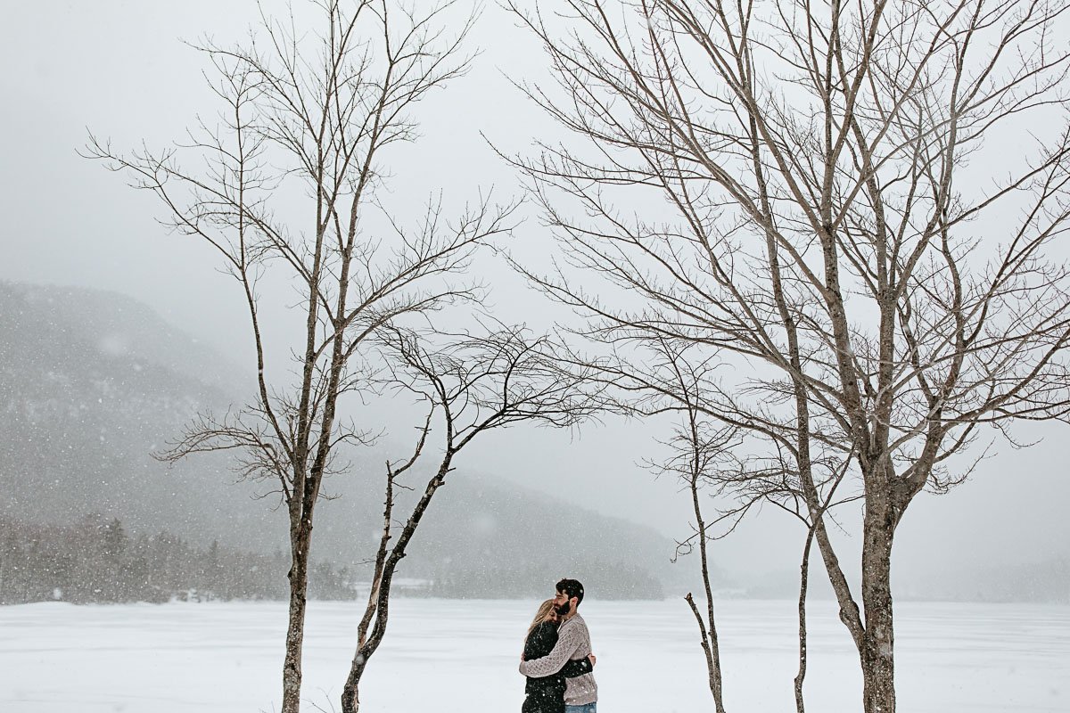  A couple wearing knit sweaters embraces under bare trees and falling snow during their engagement session at Echo Lake in New Hampshire 