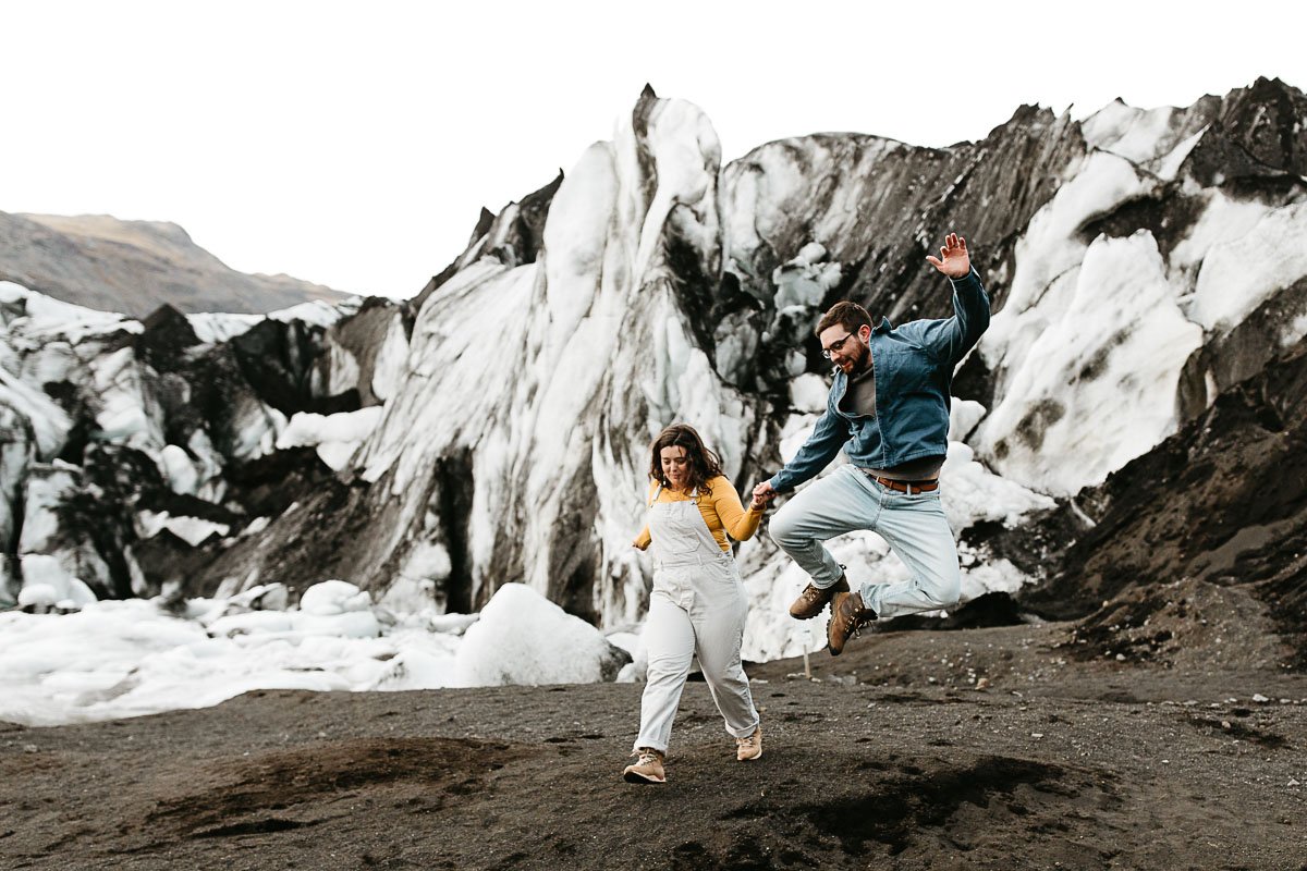  A couple skips and jumps toward the camera during their couples adventure photo session near a glacier in Iceland 