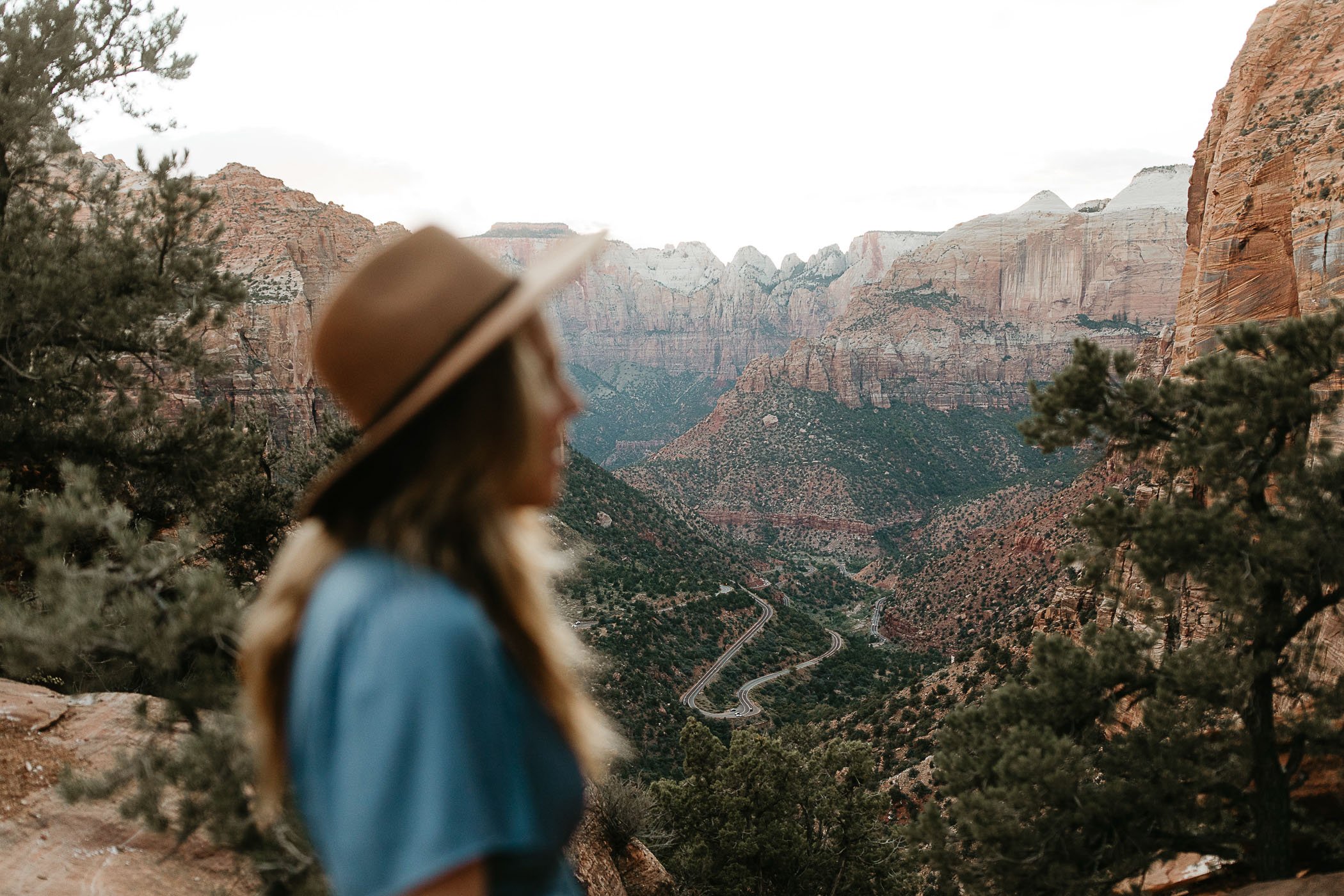  A woman in a brown wide brim hat and blue dress kisses her partner in a long grey t shirt on the Zion Canyon overlook trail. There are green trees and red rock canyons behind them. 