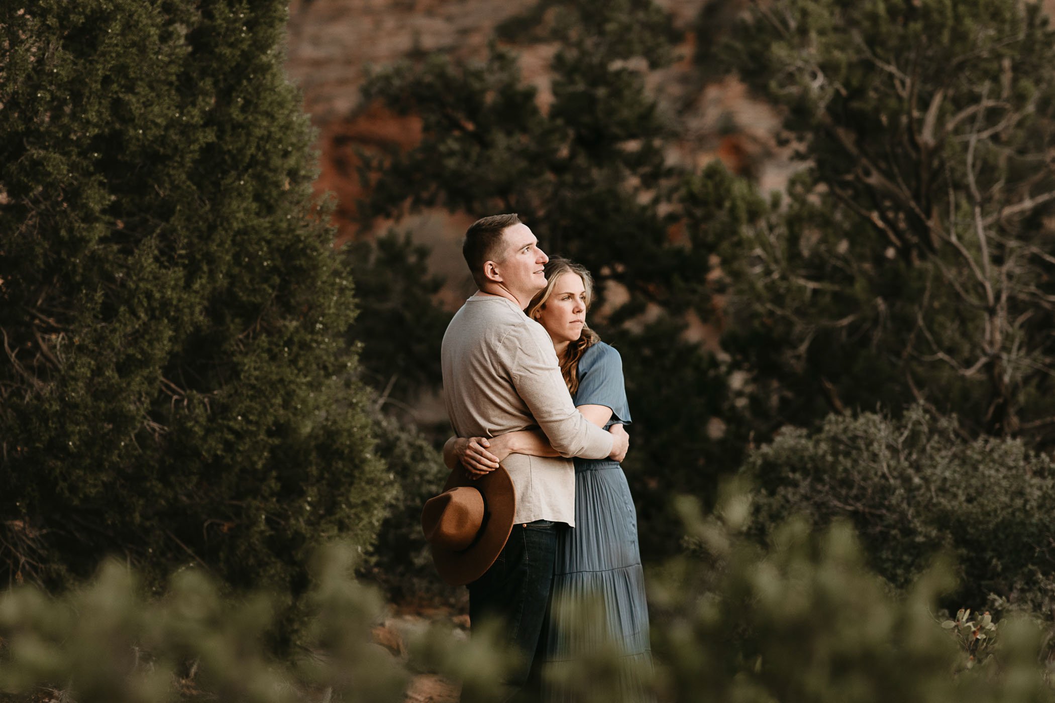  A couple in a blue dress and grey shirt embrace each other and look out toward the Canyon Overlook Trail in Zion, surrounded by pines and red rock cliffs. 