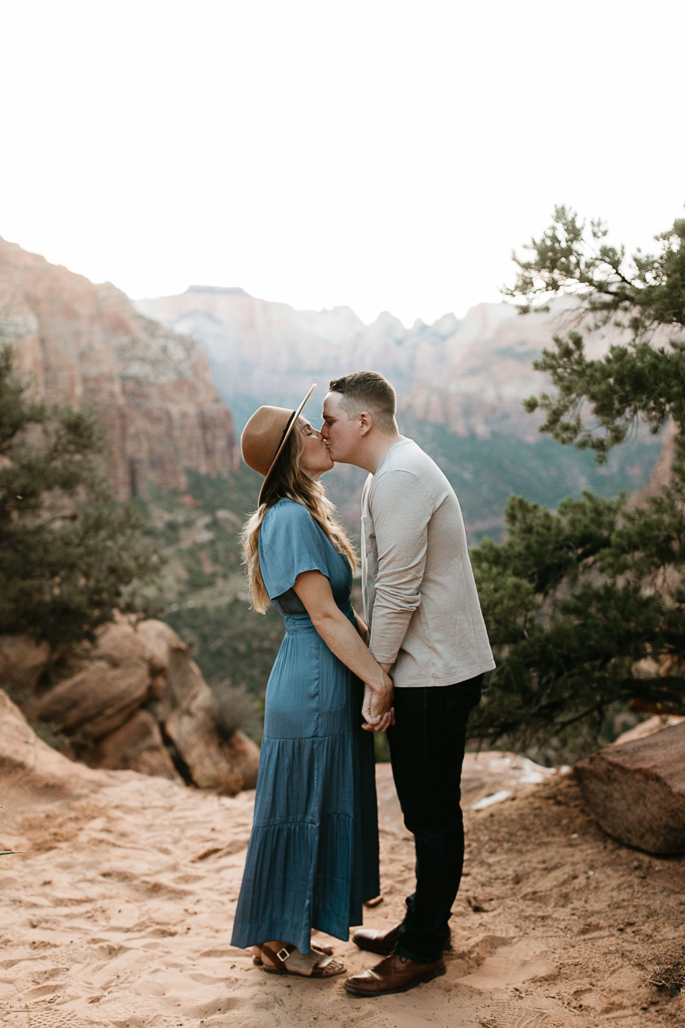  A woman in a brown wide brim hat and blue dress kisses her partner in a long grey t shirt on the Zion Canyon overlook trail. There are green trees and red rock canyons behind them. 
