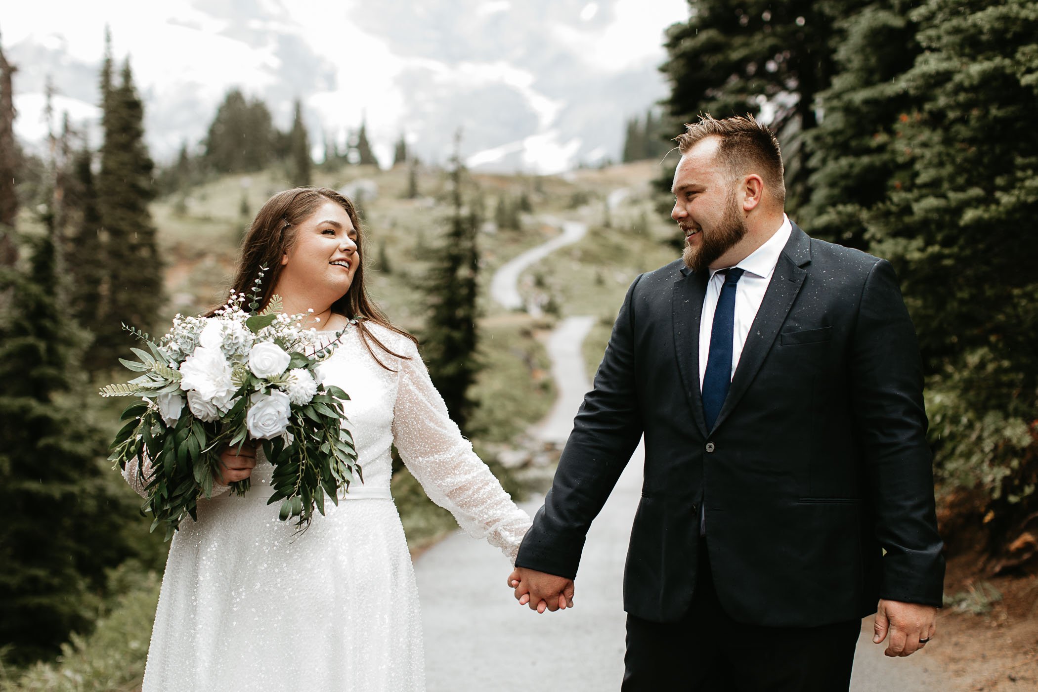  A couple in white wedding dress and suit hold hands looking at each other on a tree line path on a rainy day in Mt. Rainier National park 