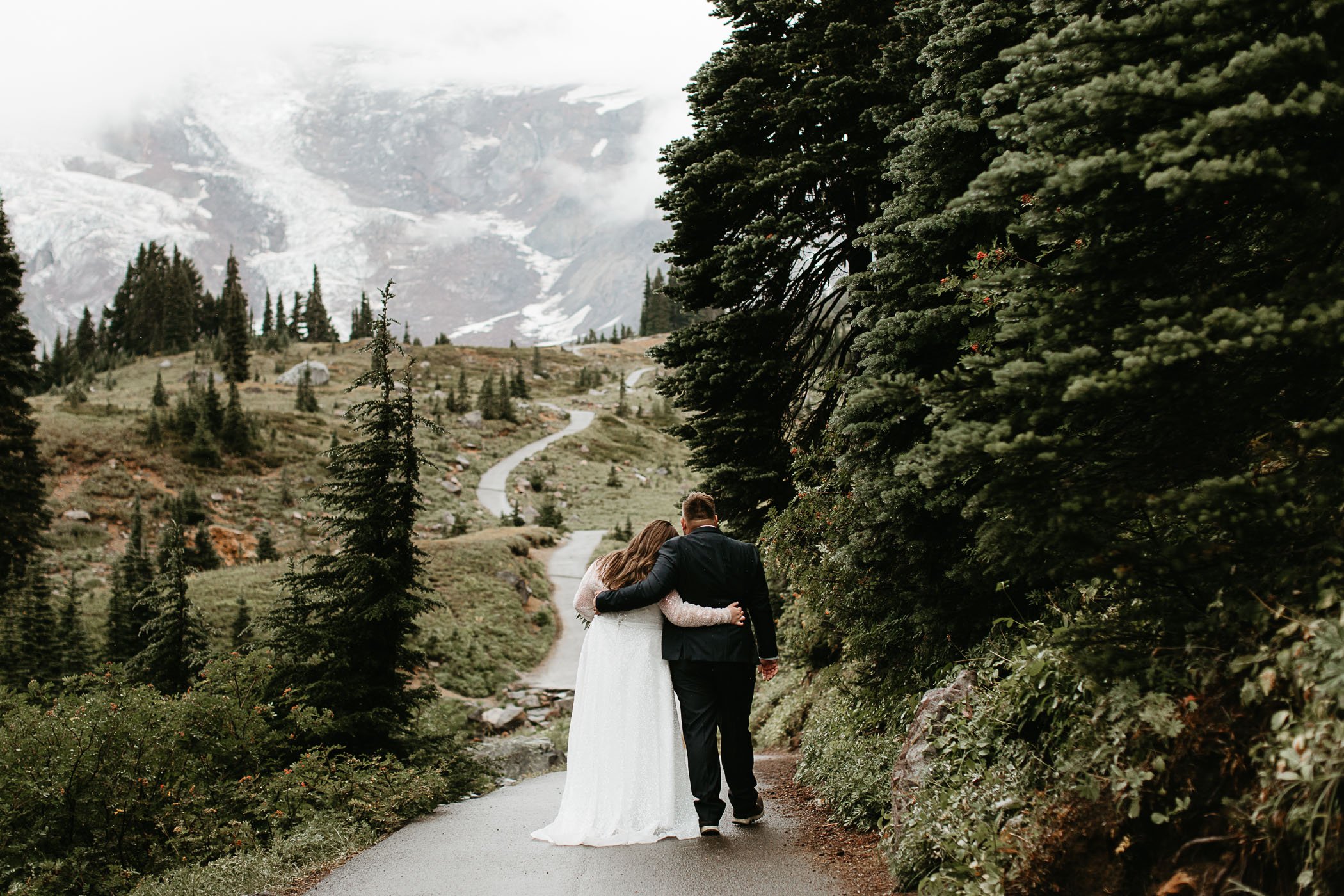  A couple walk down a wooded path in a wedding dress and suit on a rainy day in Mt. Rainier National Park. 