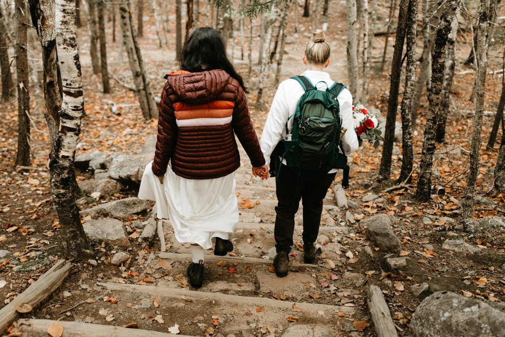  A same sex lesbian couple wearing white walk holding hands through Acadia National Park on their wedding day 