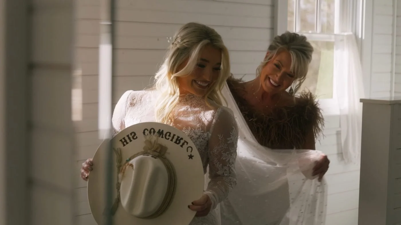 A laughing bride holding a His Cowgirl hat while her mom adjusts her veil