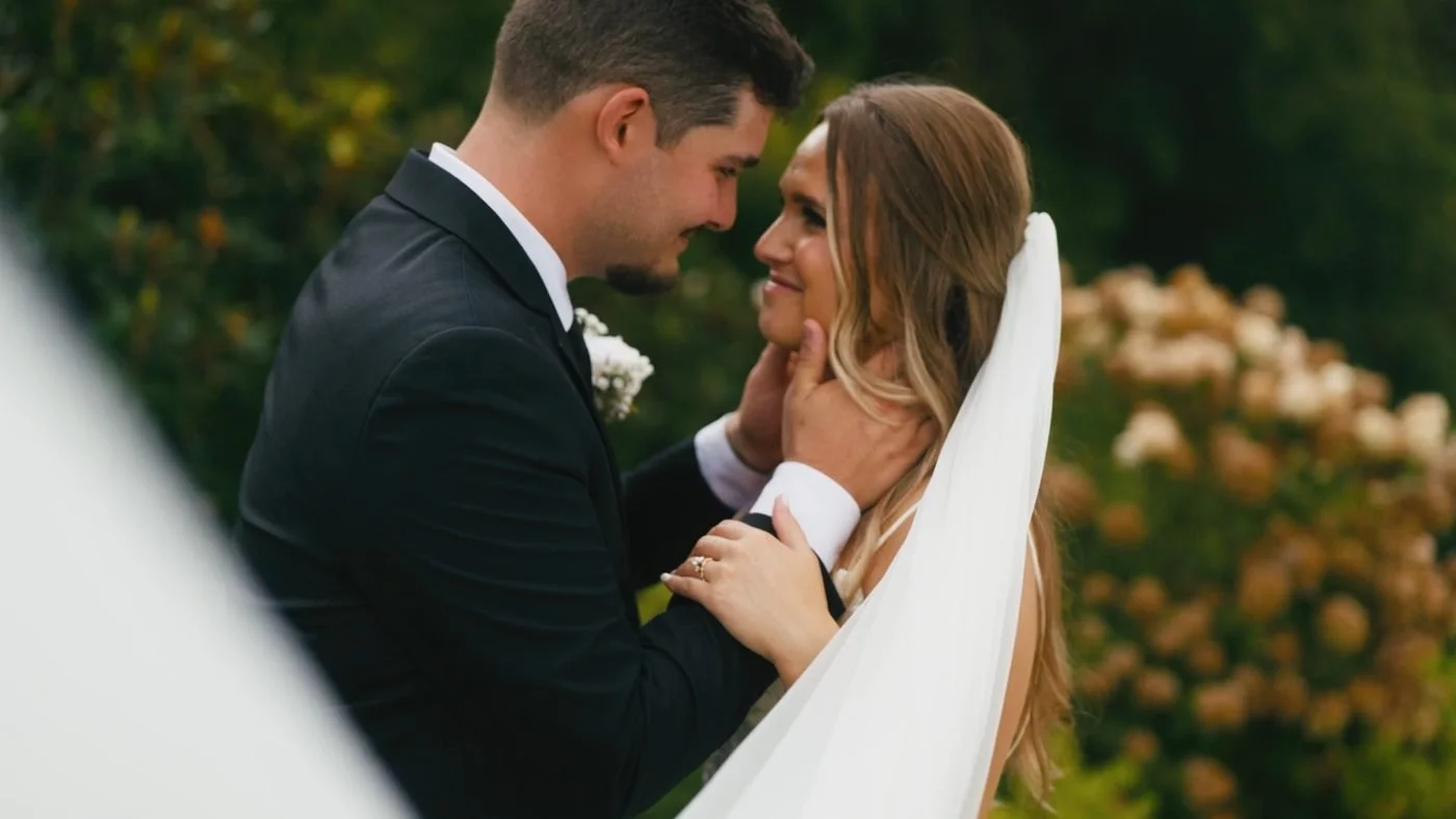 A groom holding his bride's face as she smiles, surrounded by hydrangeas