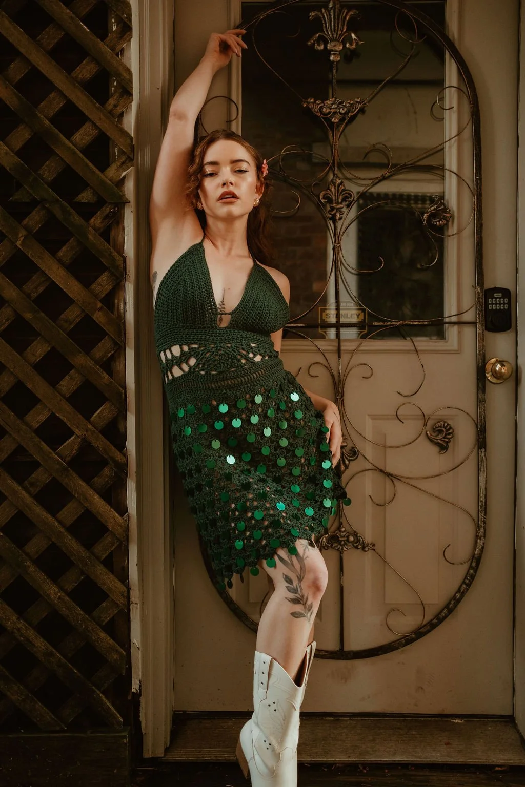 A woman with light skin and reddish-brown hair in a crochet green dress with sequins, standing with her back against a door with decorative ironwork, one arm raised above her head, and the other resting on her hip, wearing white cowboy boots.