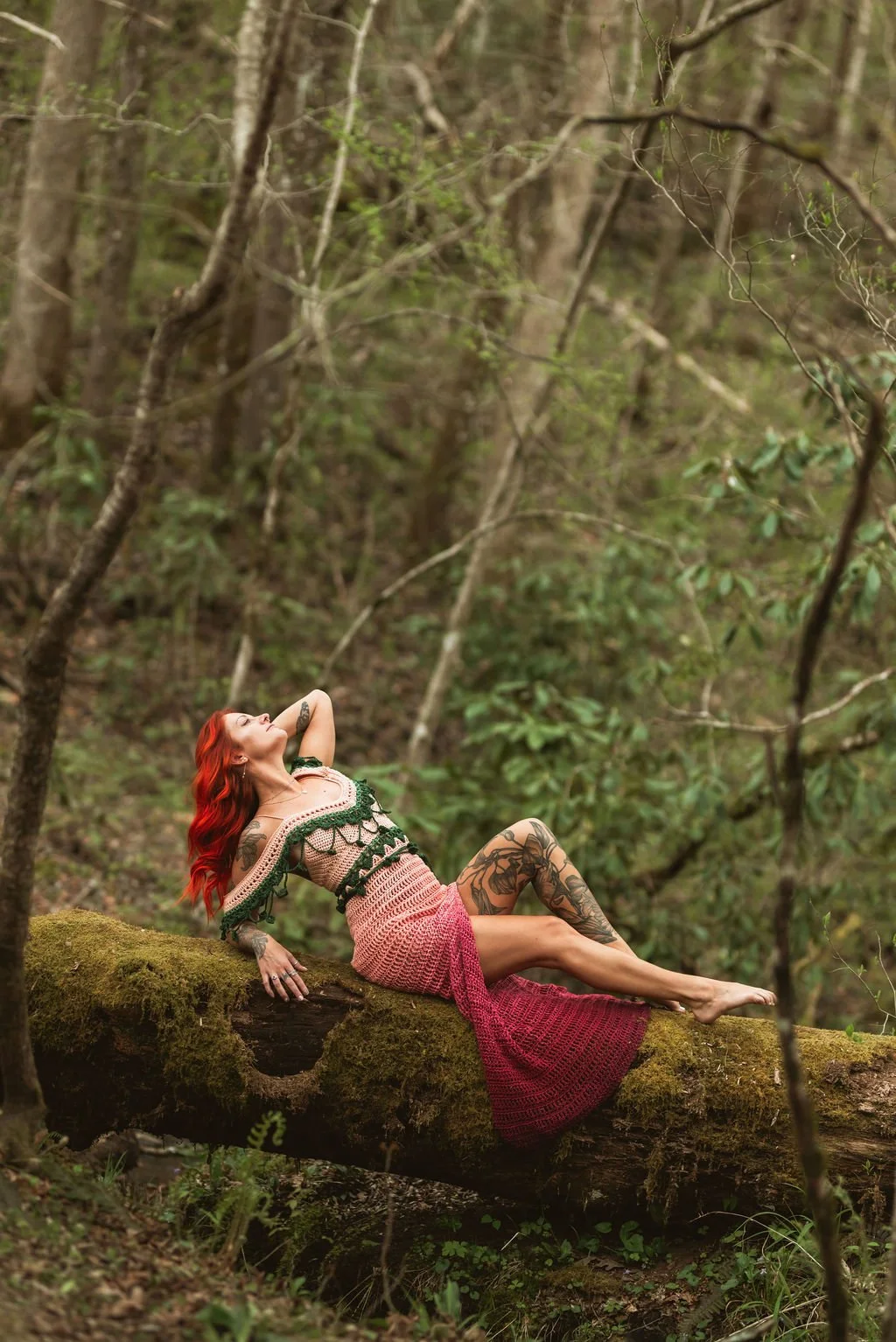 A woman with red hair and tattoos, lying on a moss-covered fallen tree in a forest, wearing a crochet dress with pink and green accents.