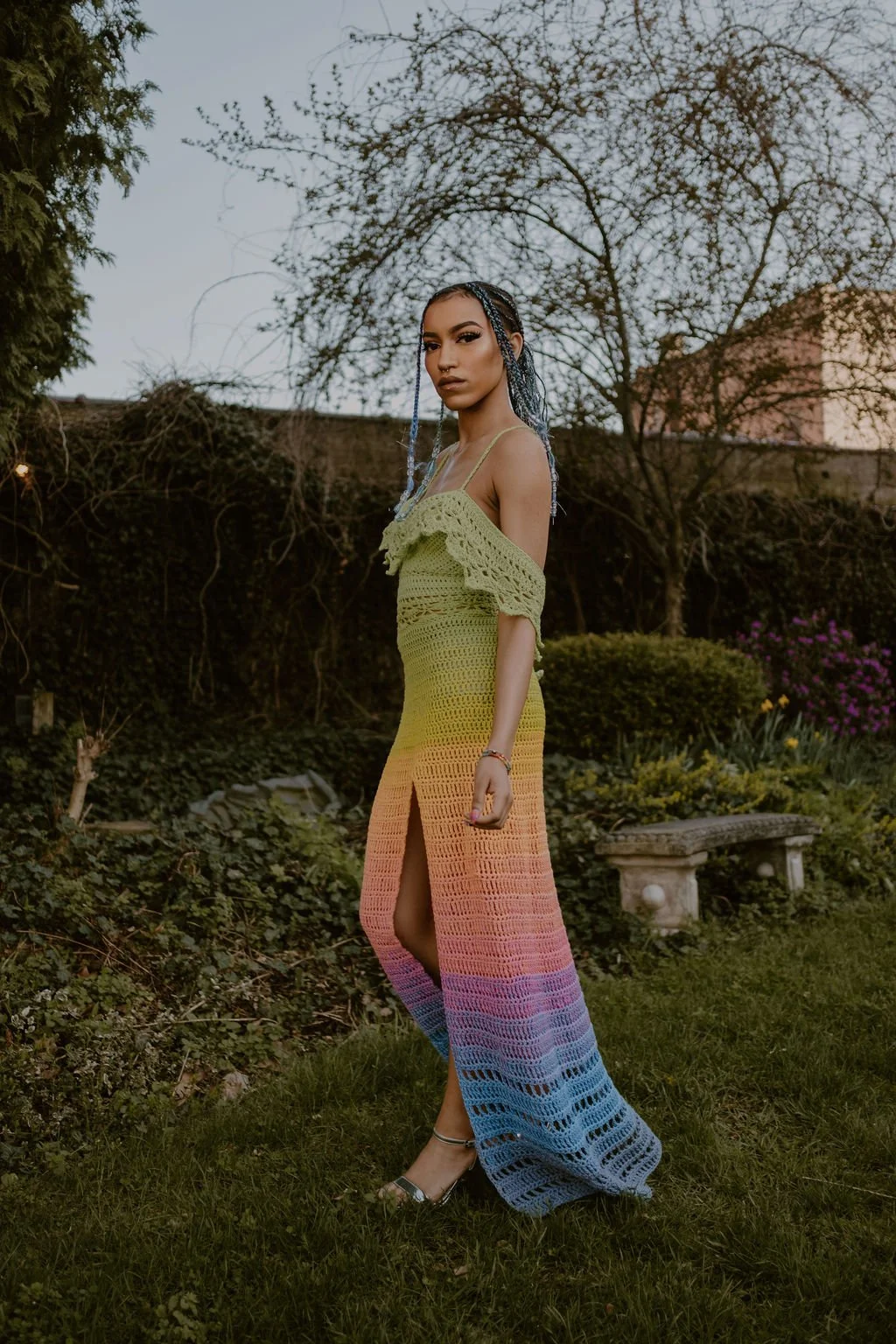 Young woman with braided hair in a colorful crochet dress standing in a garden during dusk, with trees and shrubs in the background.