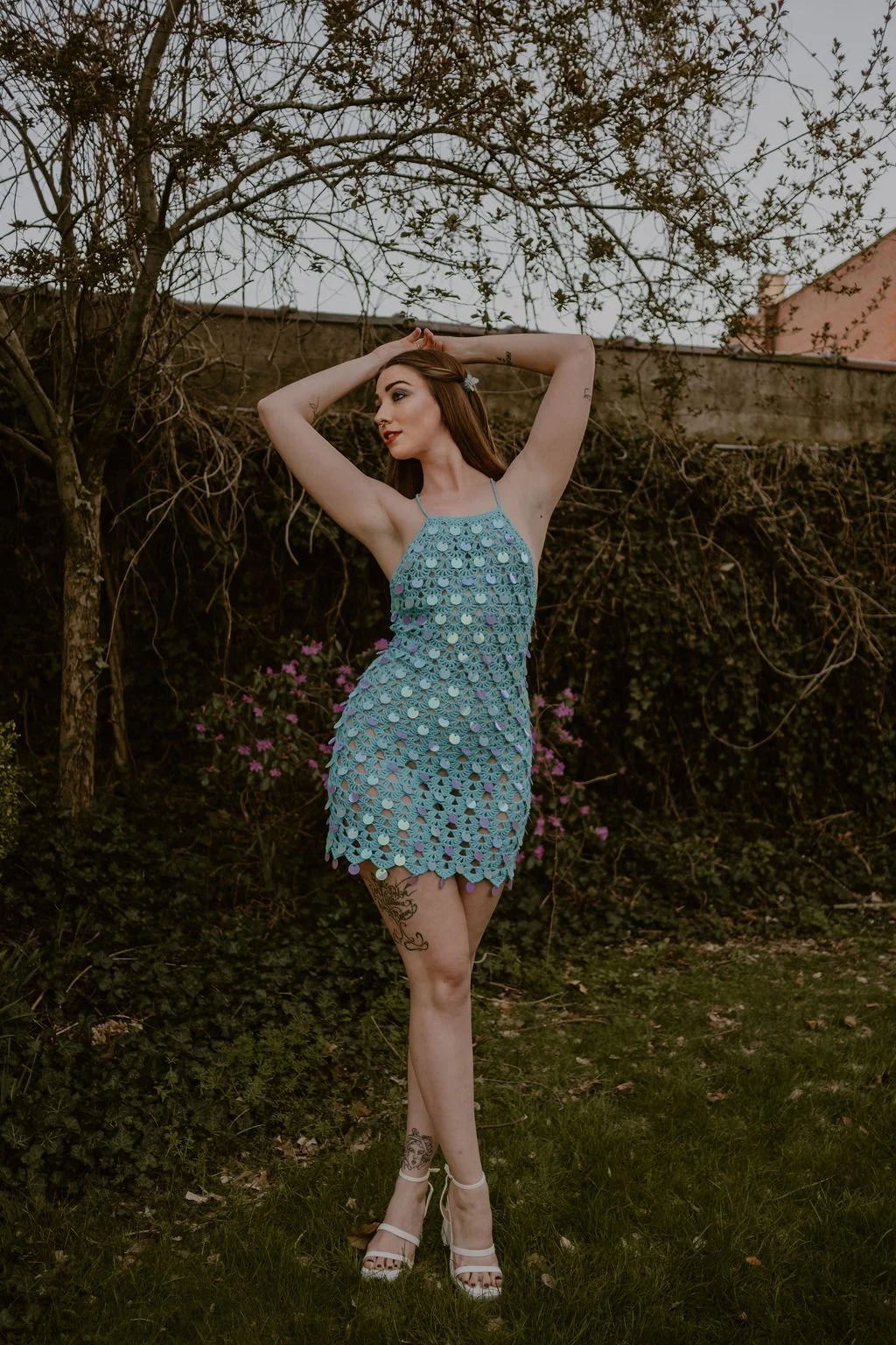 A woman in a blue crochet dress and white high heels standing outdoors with her arms raised above her head, surrounded by trees and pink flowers.