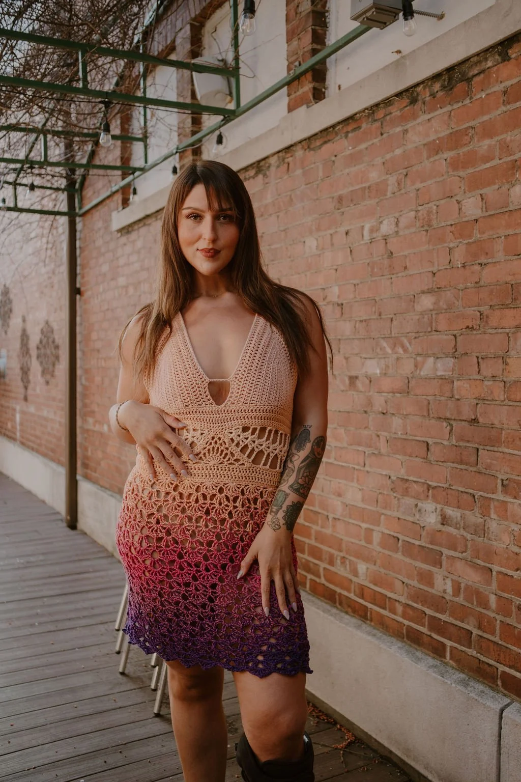 A woman with long brown hair and tattoos on her left arm is standing on a wooden deck beside a brick wall, wearing a sleeveless crochet dress with a gradient pattern from beige to purple.