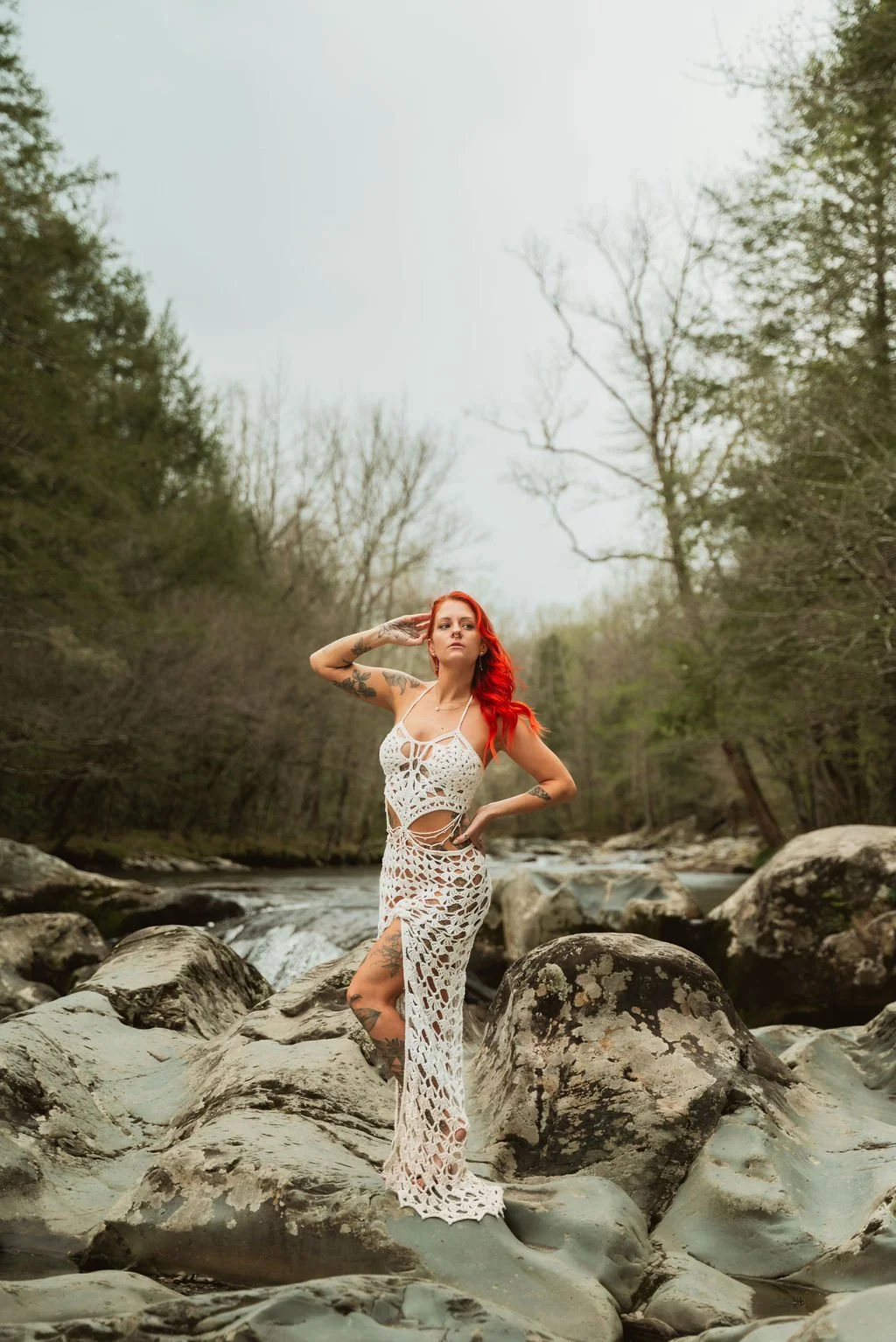 A woman with red hair wearing a white woven dress poses on rocks in a creek surrounded by trees.