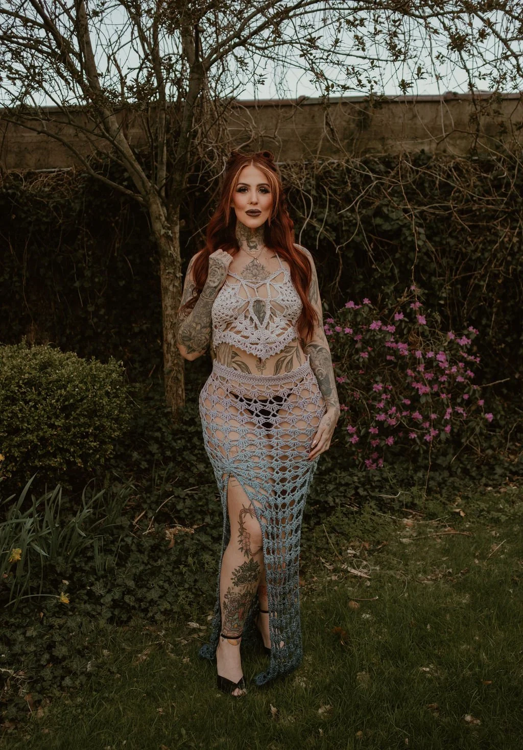 A woman with long red hair standing outdoors in front of a tree and pink flowers, wearing a crochet dress and high heels.