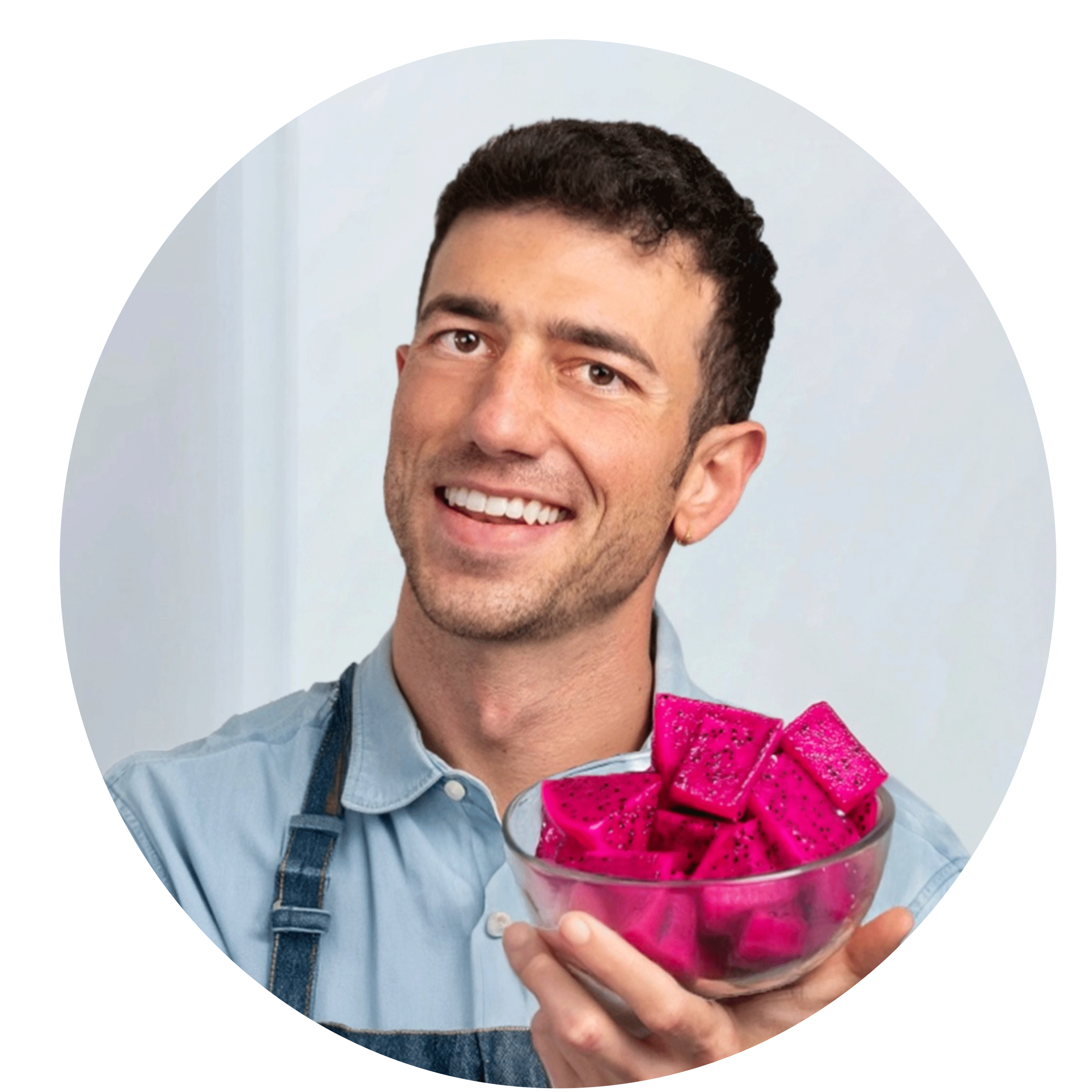 A chef holding a large glass cup of bright pink dragon fruit. The man is TikTok chef Mario Fabbri also known as @veganrecipes, Mario is smiling and wearing a light blue chef apron.