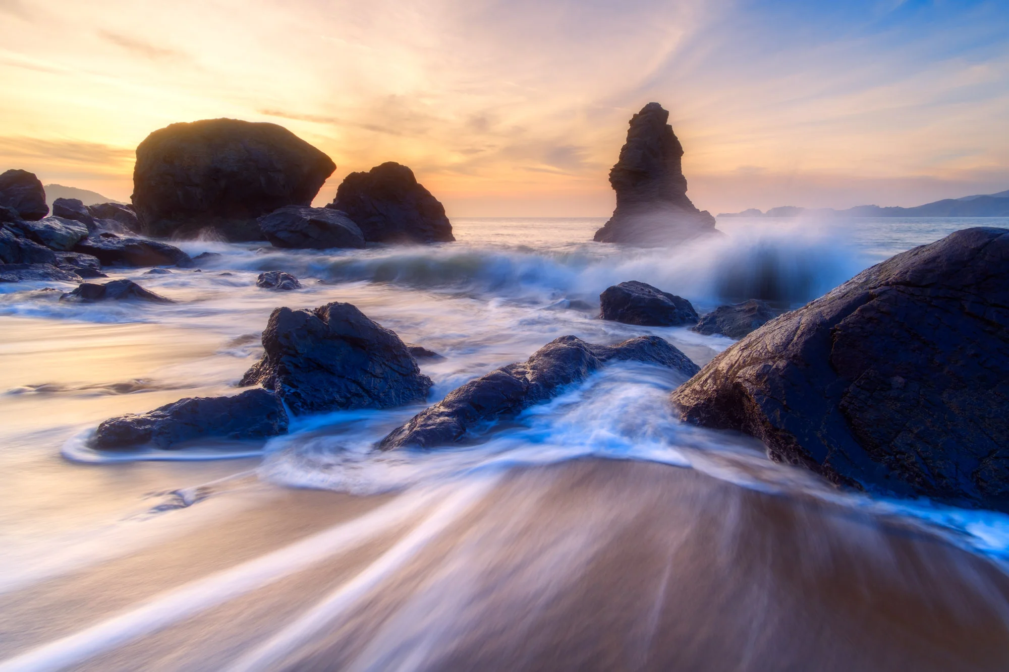 As sea water from one washes back out, the next wave crashes into the rocks at sunset