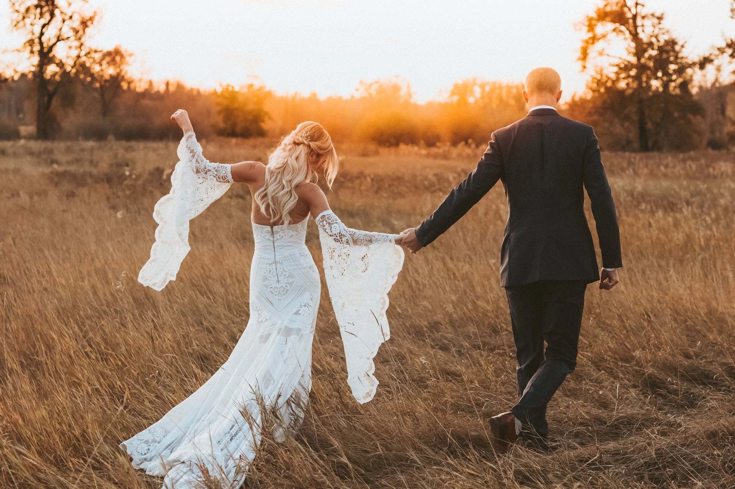 Bride and groom walking hand in hand through a golden field at sunset during an intimate wedding in Fish Creek Park, Calgary, photographed by Maxine LaGarde Photography.