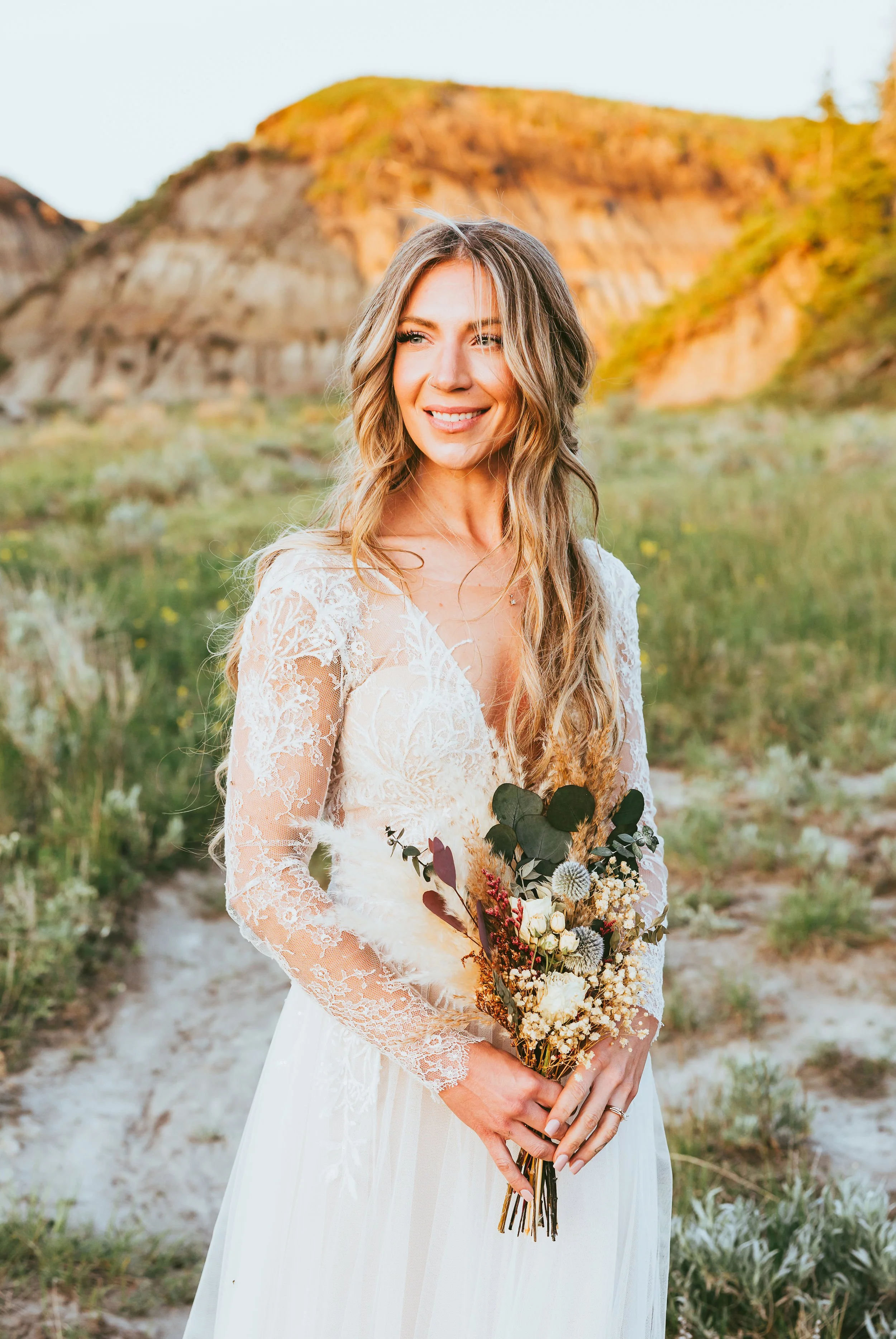 Bride standing in flowing lace dress holding bouquet in Drumheller Badlands landscape at sunset in Alberta Canada