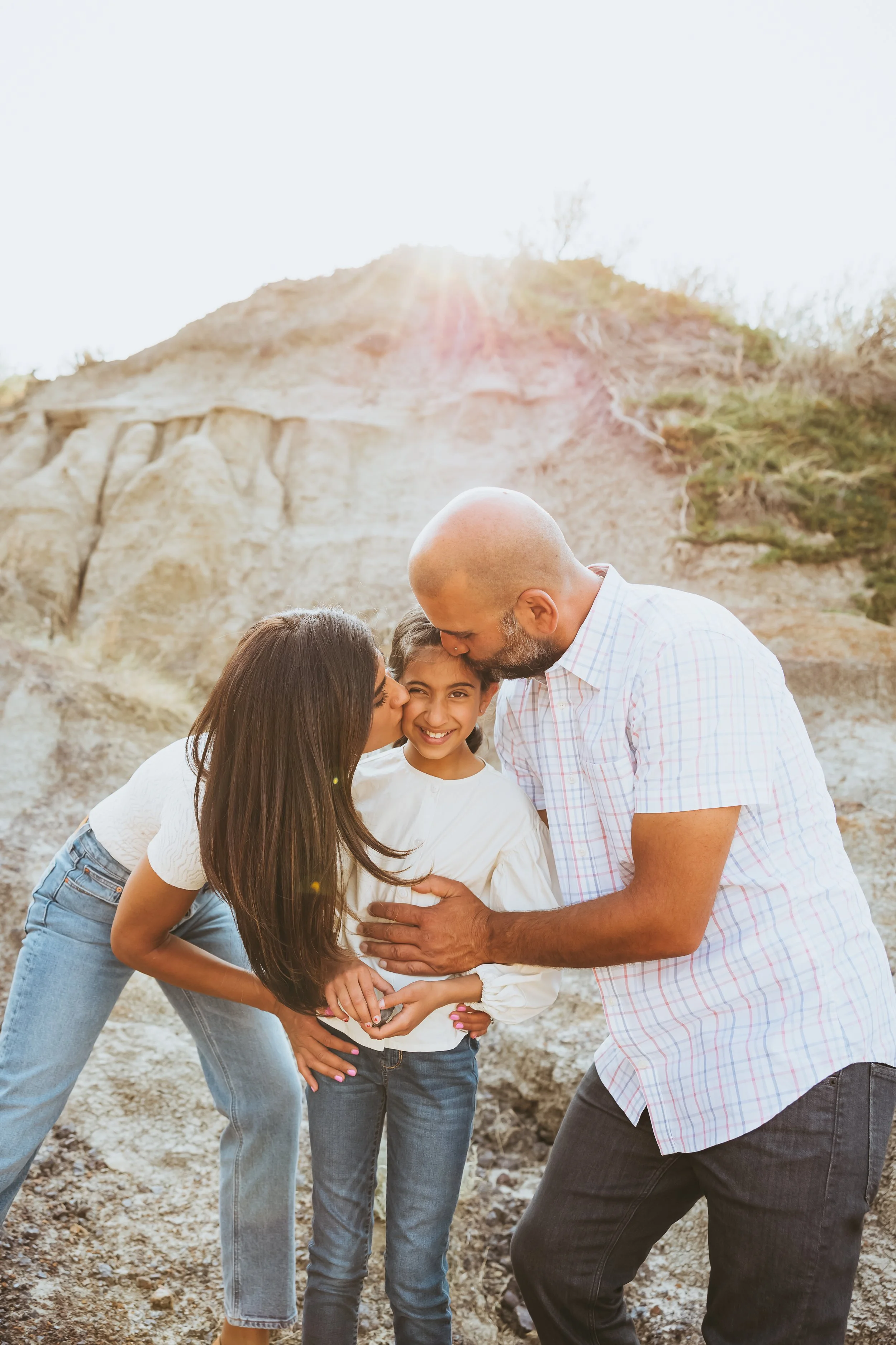 Candid family photo in the Alberta Badlands with parents kissing their child during a warm sunset session