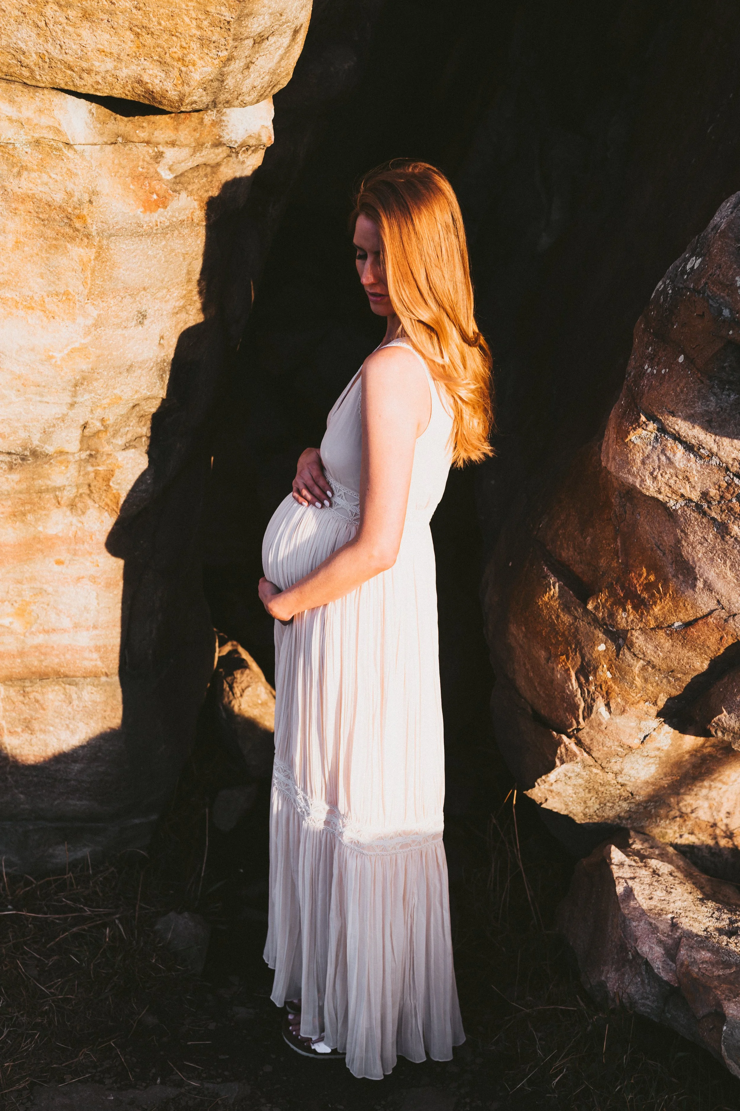 Pregnant woman in a flowing cream dress standing between sandstone rock formations during a golden hour maternity photoshoot in the Alberta Badlands