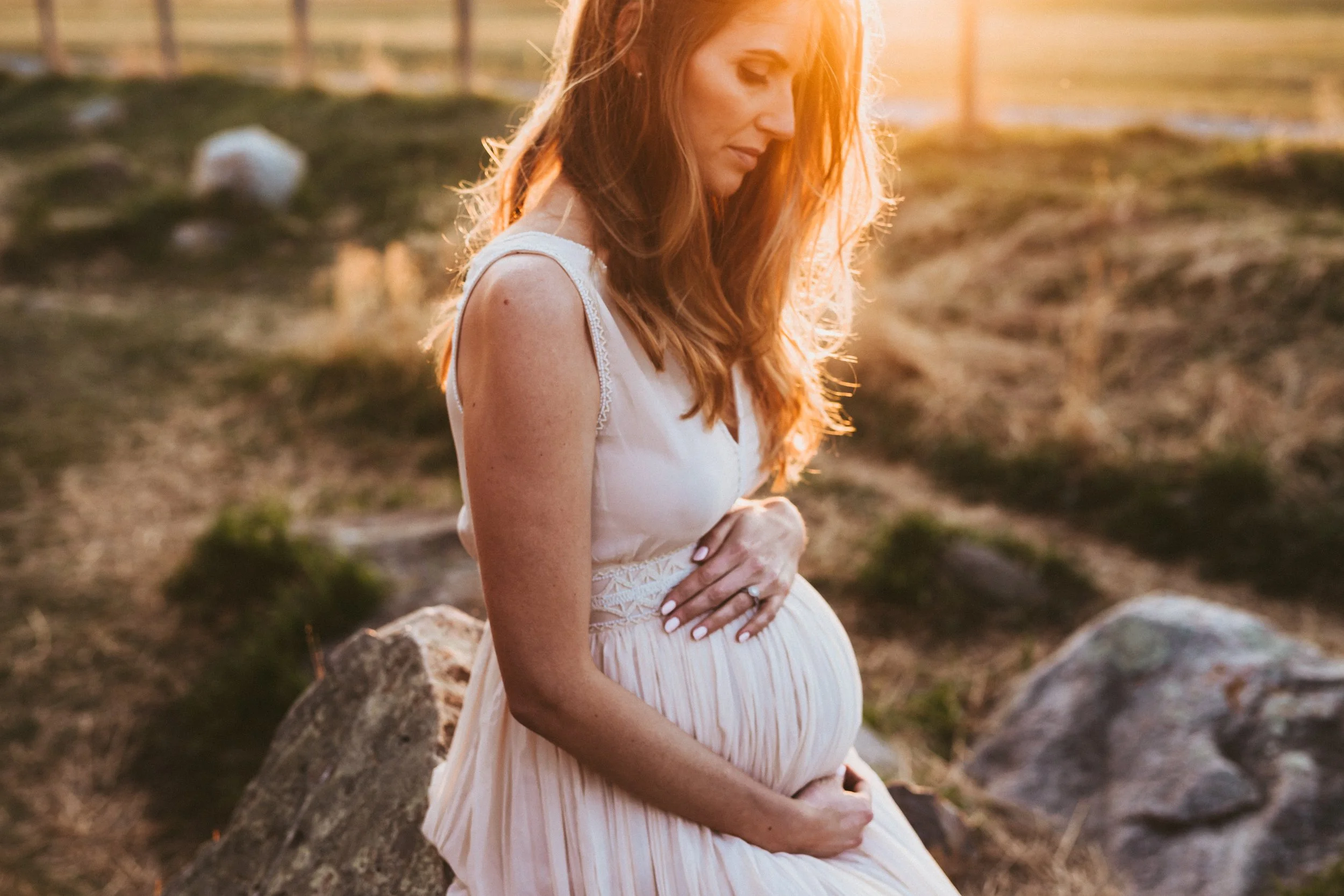 Pregnant woman sitting on a rock in warm sunset light, wearing a flowing neutral dress during an outdoor maternity photoshoot