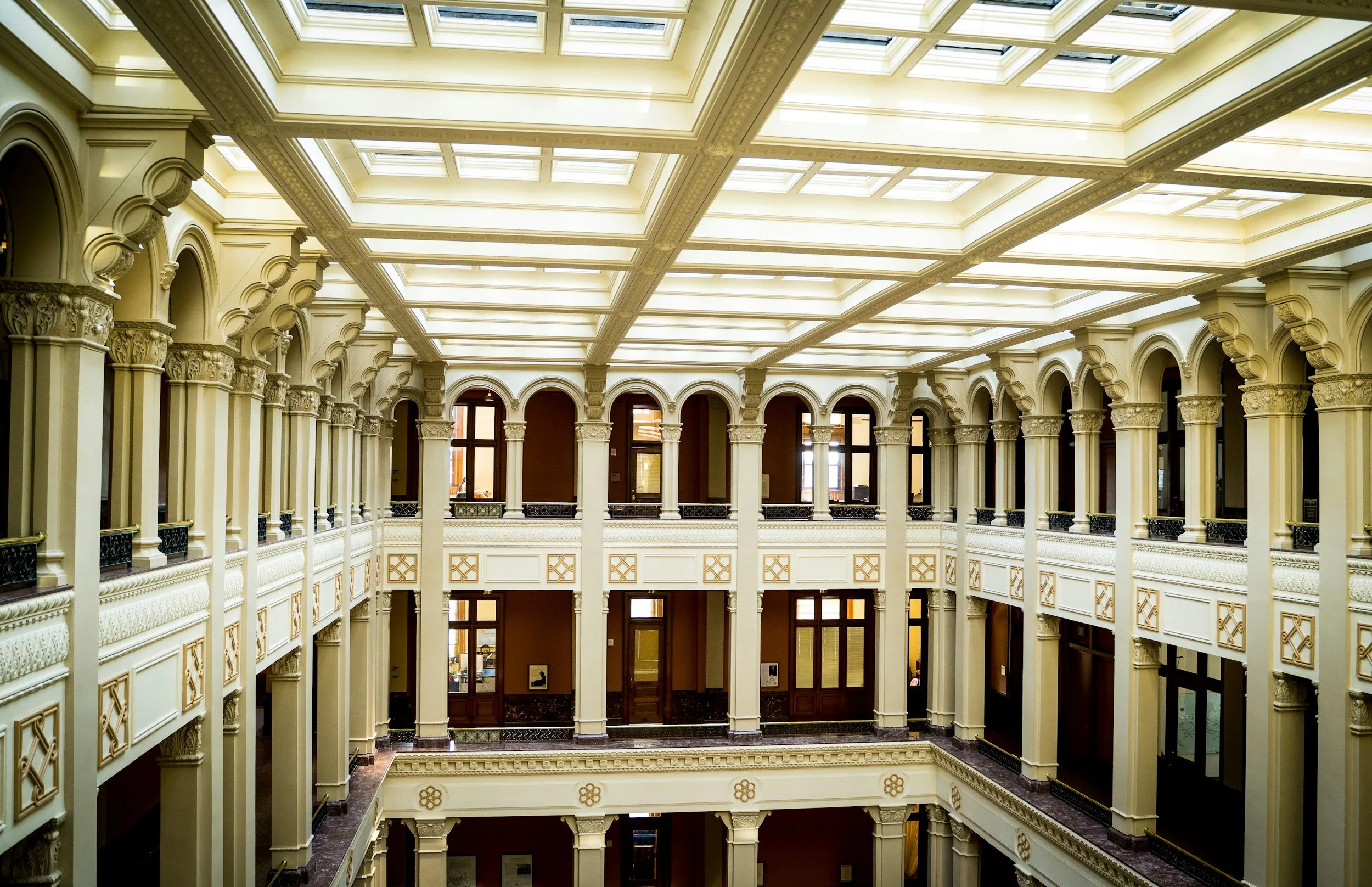Interior view of a grand, multi-story building with ornate architecture, arches, columns, and a large skylight ceiling.