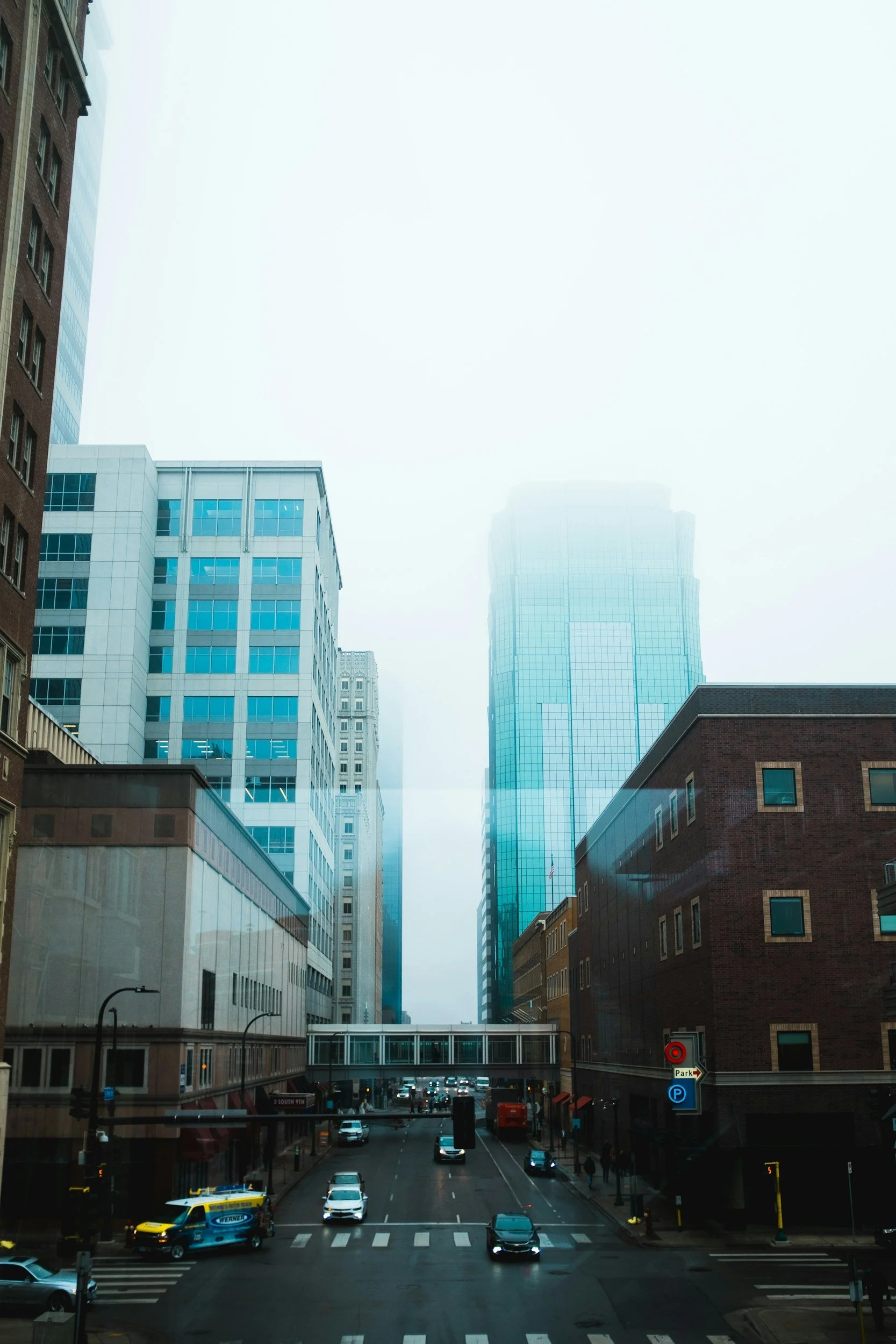 City street with tall modern buildings, some with glass facades, and fog enveloping the upper parts of the buildings.