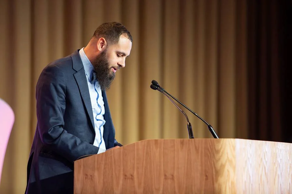 Dustin pictured standing at a podium looking down as he reads his speech.