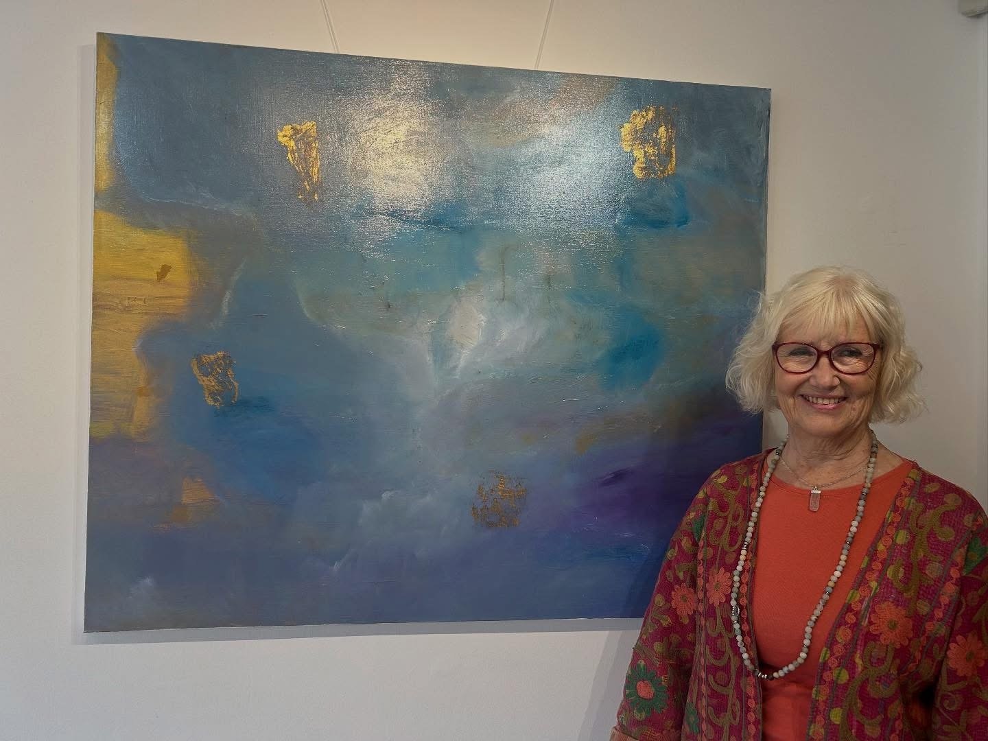 An elderly woman with glasses and curly gray hair smiling standing in front of a large abstract painting with blue, gold, and purple colors.