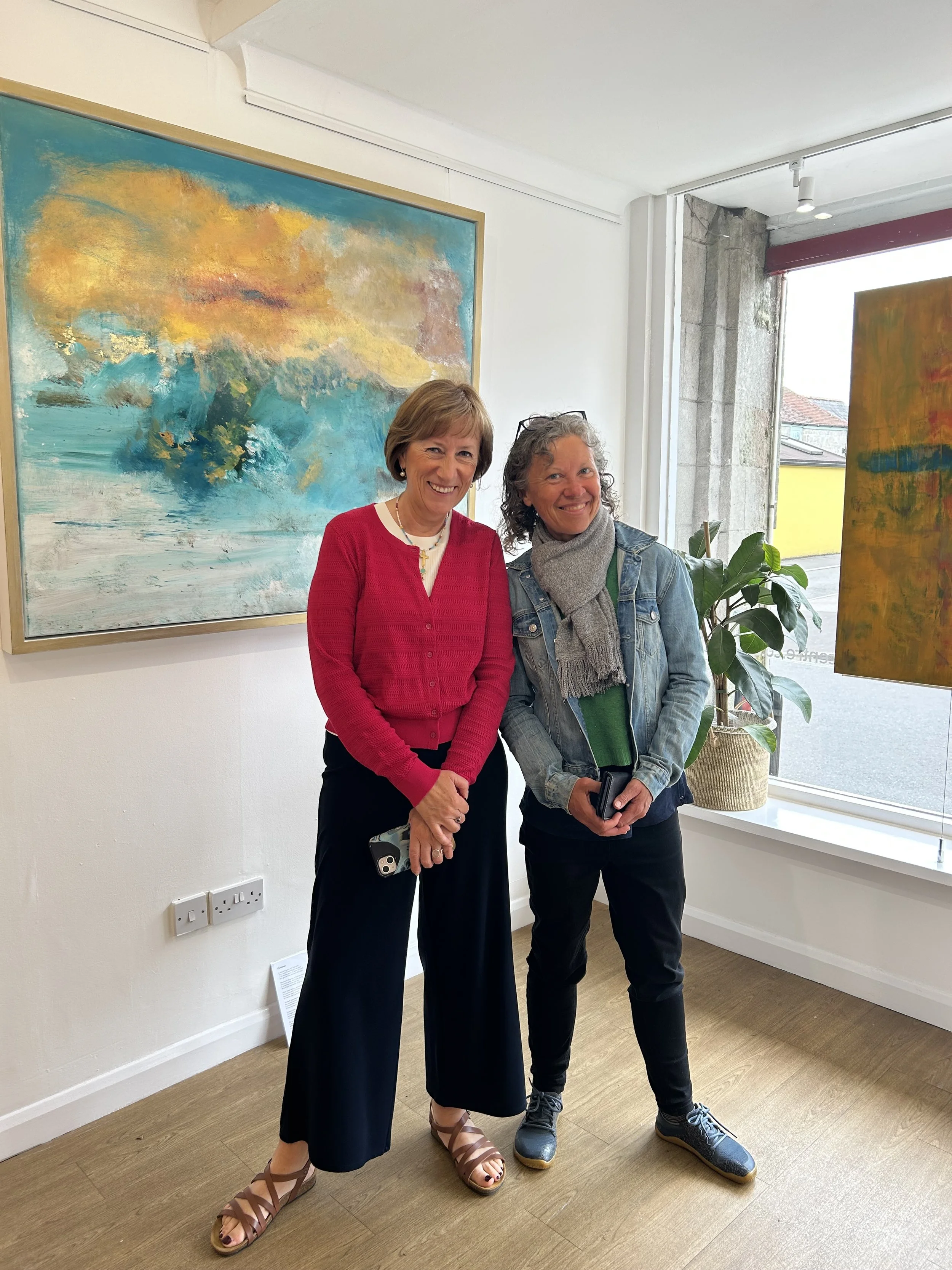 Two women standing in an art gallery with abstract paintings on the wall and a potted plant near a large window.
