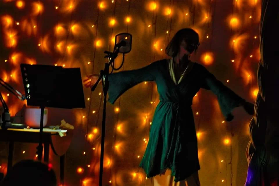 A woman in a teal dress and sunglasses standing with outstretched arms in front of a wall decorated with orange string lights, with a music stand and microphone nearby.