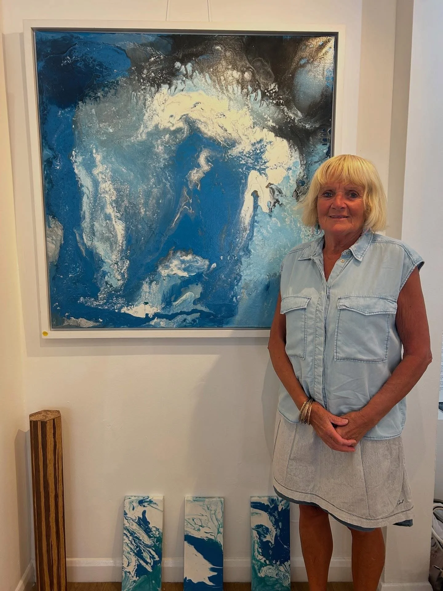 An older woman with blonde hair wearing a sleeveless light blue shirt and a beige skirt stands beside a large abstract painting in shades of blue, white, and black, hanging on a white wall. There are four smaller pieces of similar abstract art on the